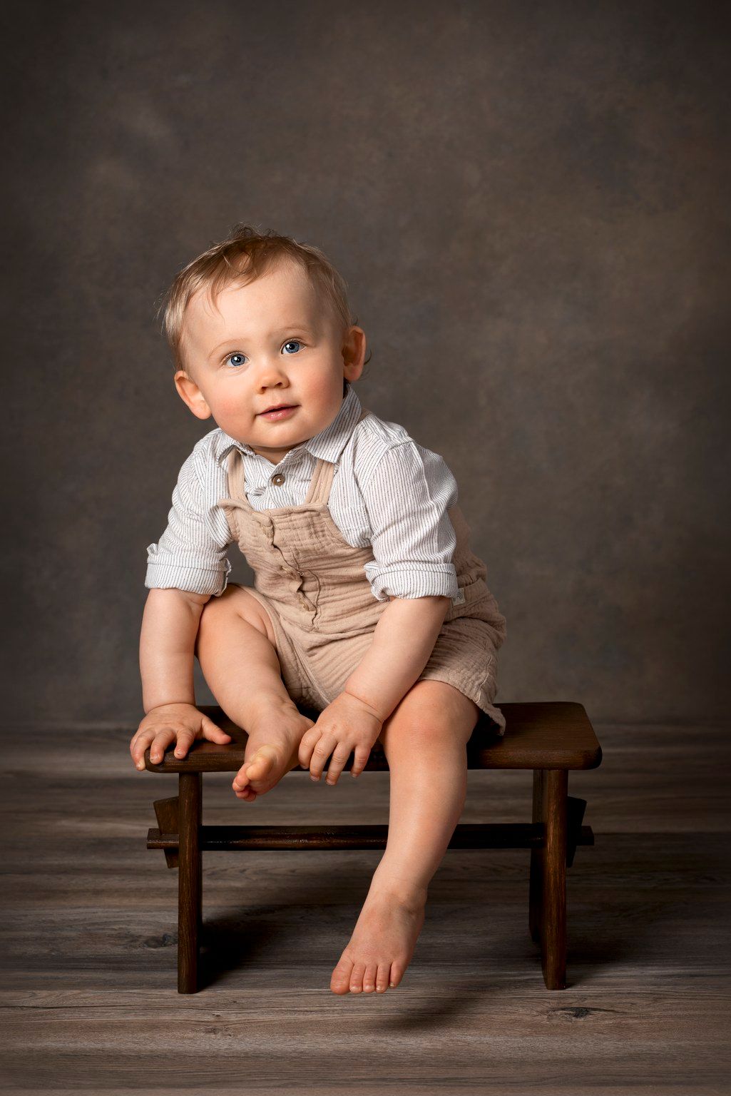 Adorable baby boy in beige overalls and white shirt sitting on a wooden stool against a gray studio backdrop.