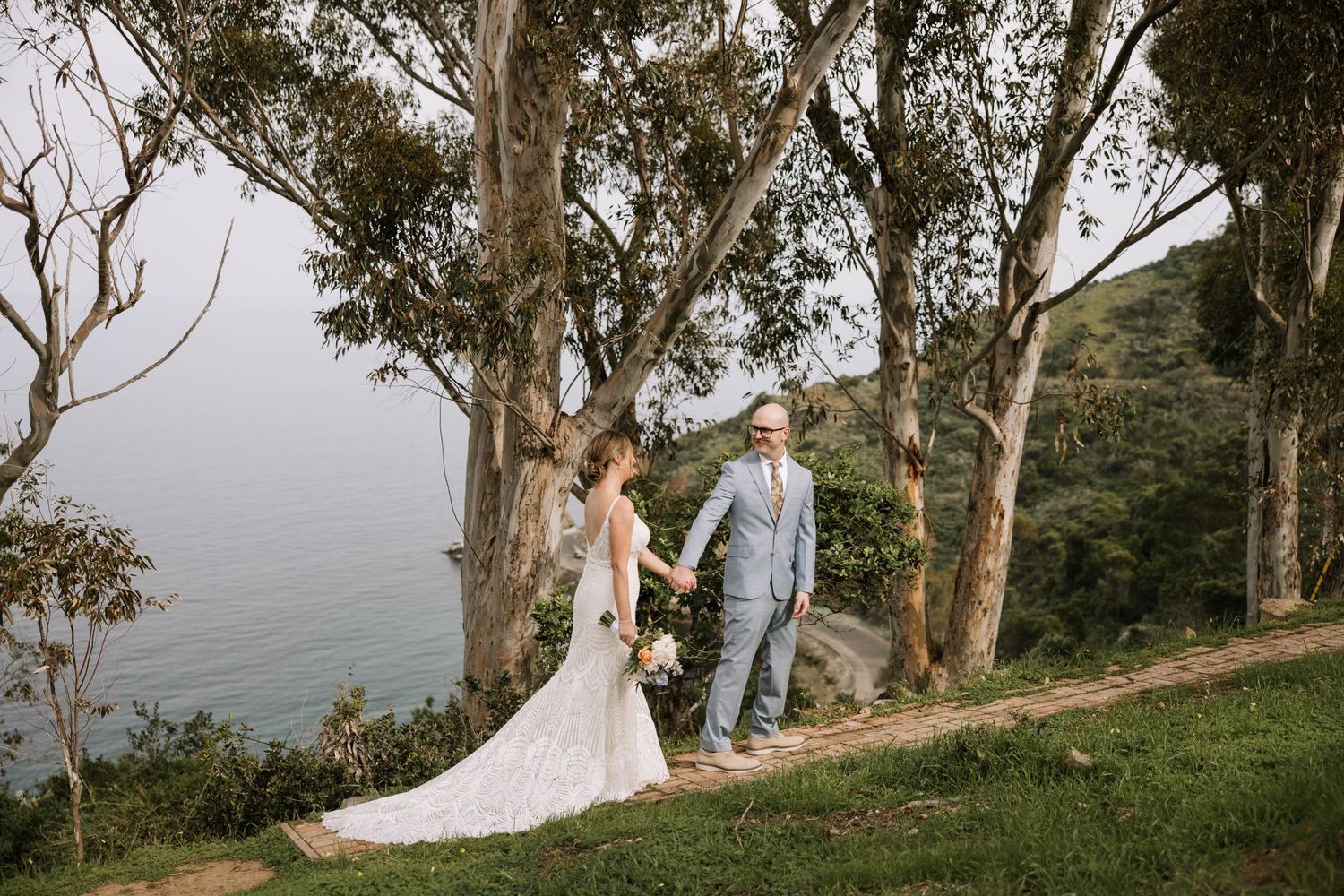 Bride and groom holding hands on scenic coastal hillside path surrounded by eucalyptus trees.