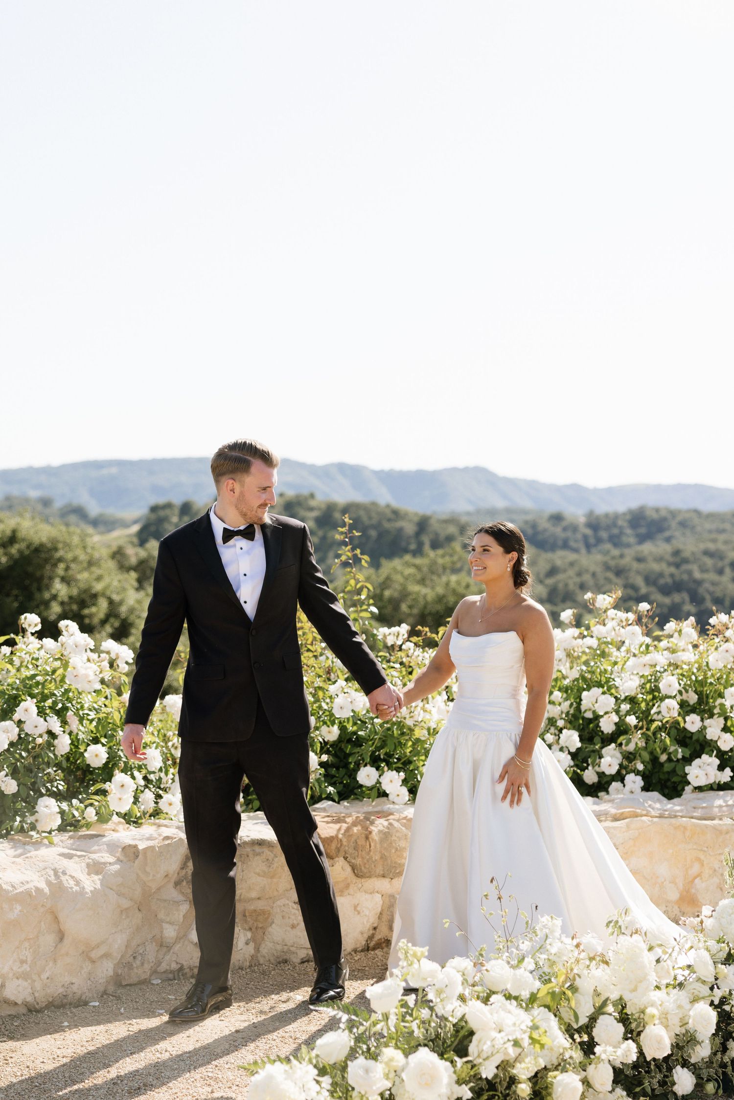 Bride and groom hold hands among white roses with mountain views on their wedding day.
