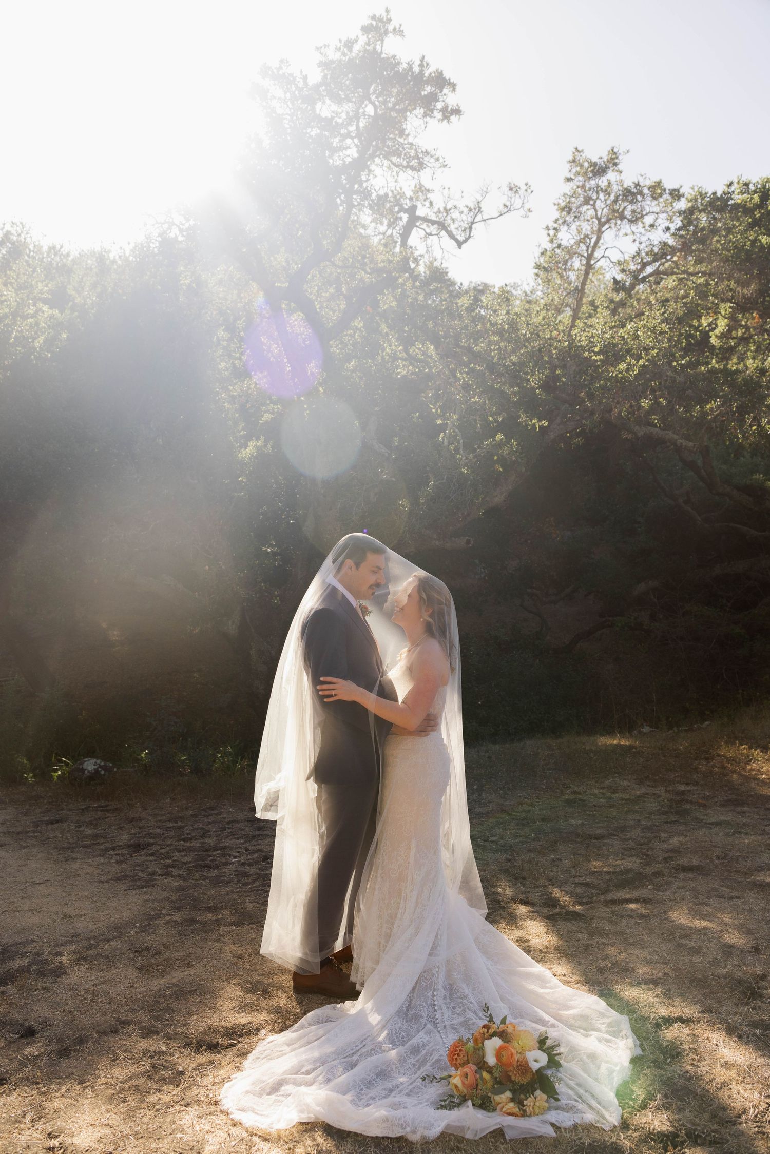 Bride and groom sharing a kiss under lace veil in sunlit forest clearing with bridal bouquet.