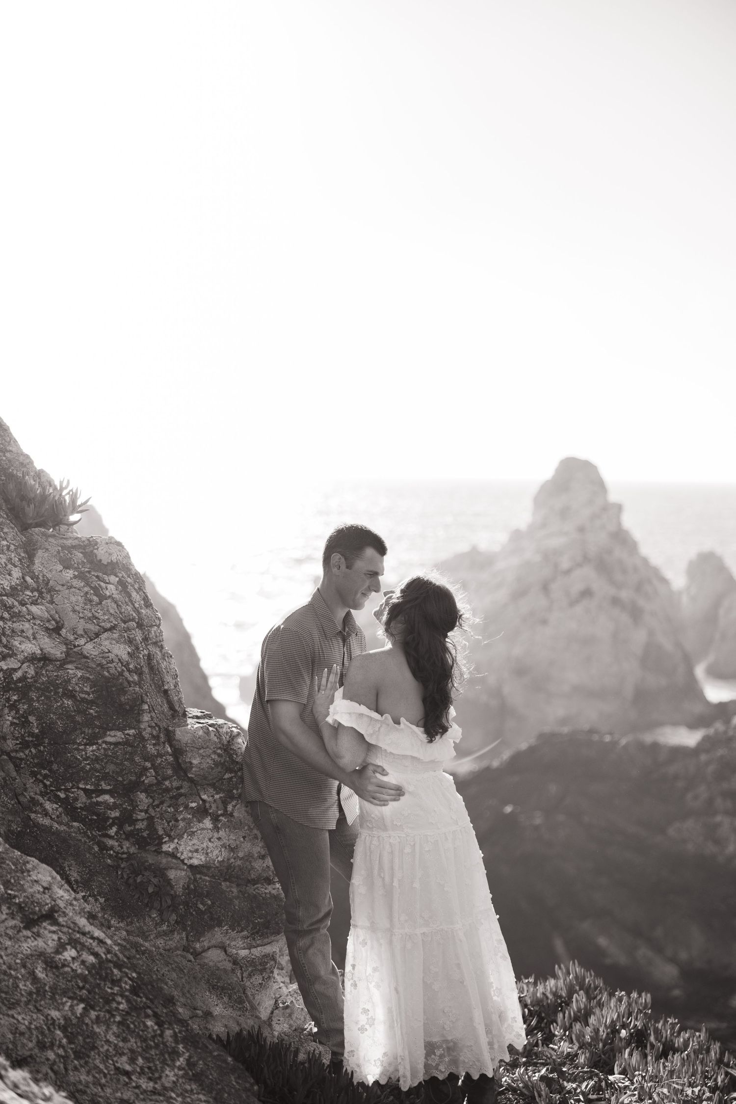 Black and white photo of a romantic couple embracing on a rocky coastal cliff with ocean and mountains in the background.