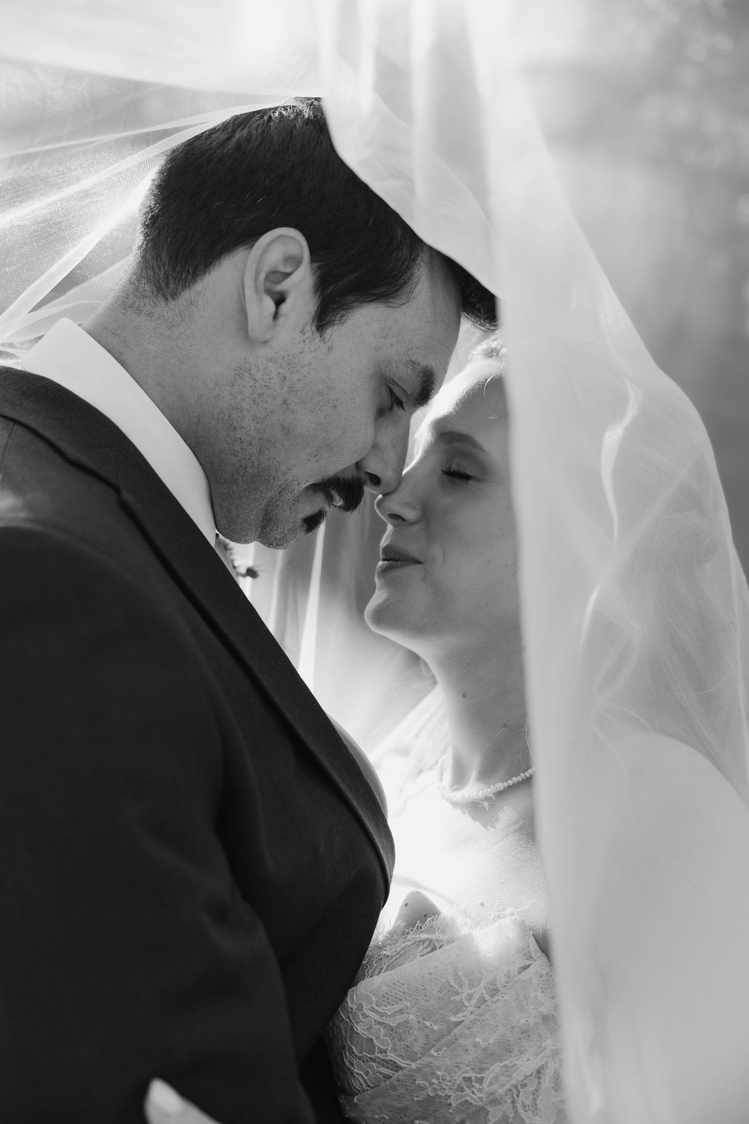 Black and white romantic wedding photo of bride and groom nearly kissing under flowing bridal veil.
