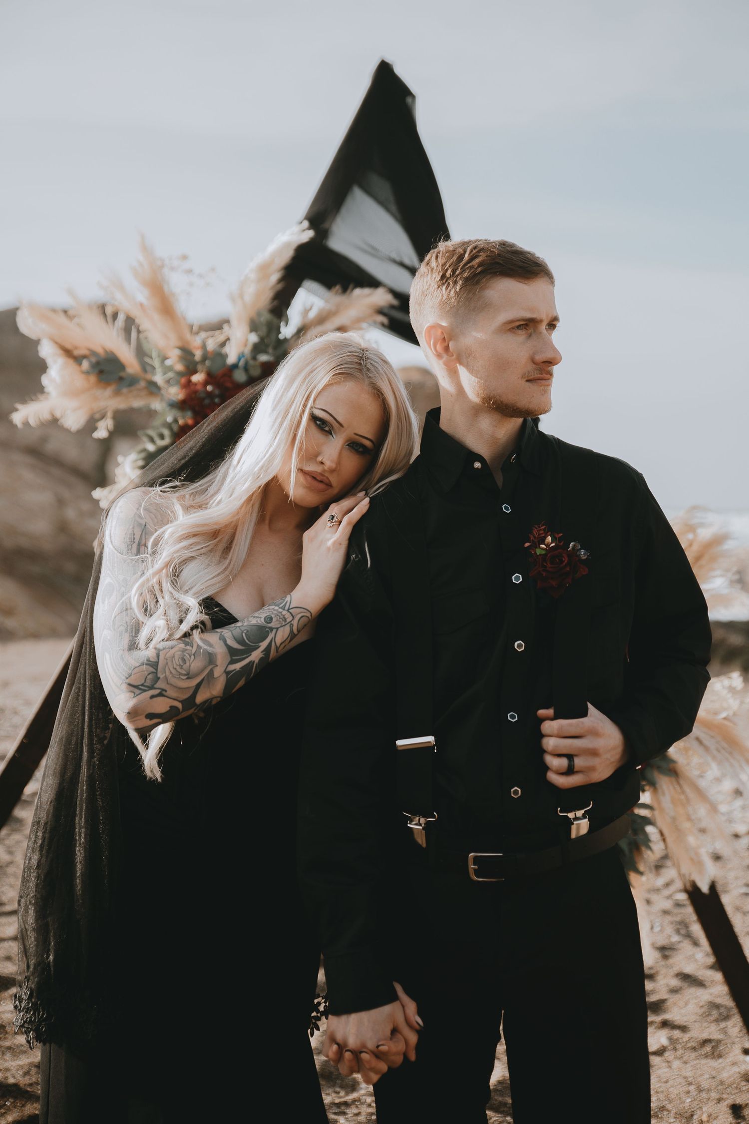 Gothic wedding couple in all-black attire holding hands before dark boho arch on sandy beach. Montana De Oro beach, California