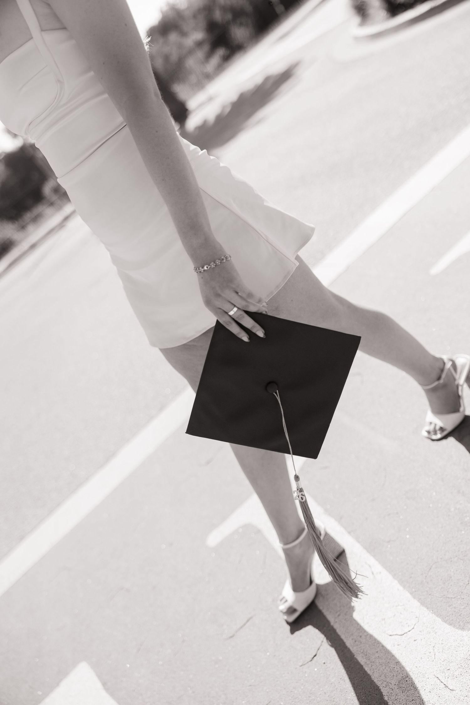 Black and white photo of a graduate in white dress and heels holding a mortarboard cap on a sunny street.