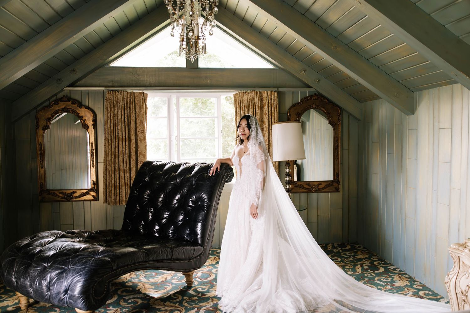 Bride in flowing white gown with long veil standing in rustic room with chandelier and vintage mirrors.