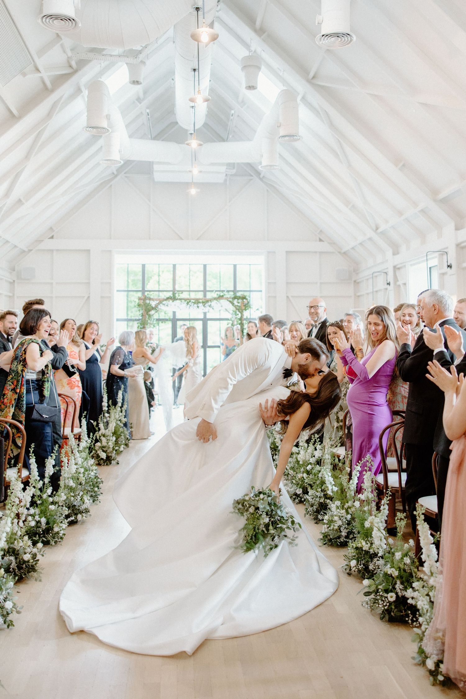 Bride and groom share a romantic dip kiss in a bright white chapel aisle lined with lush greenery during their wedding ceremony.