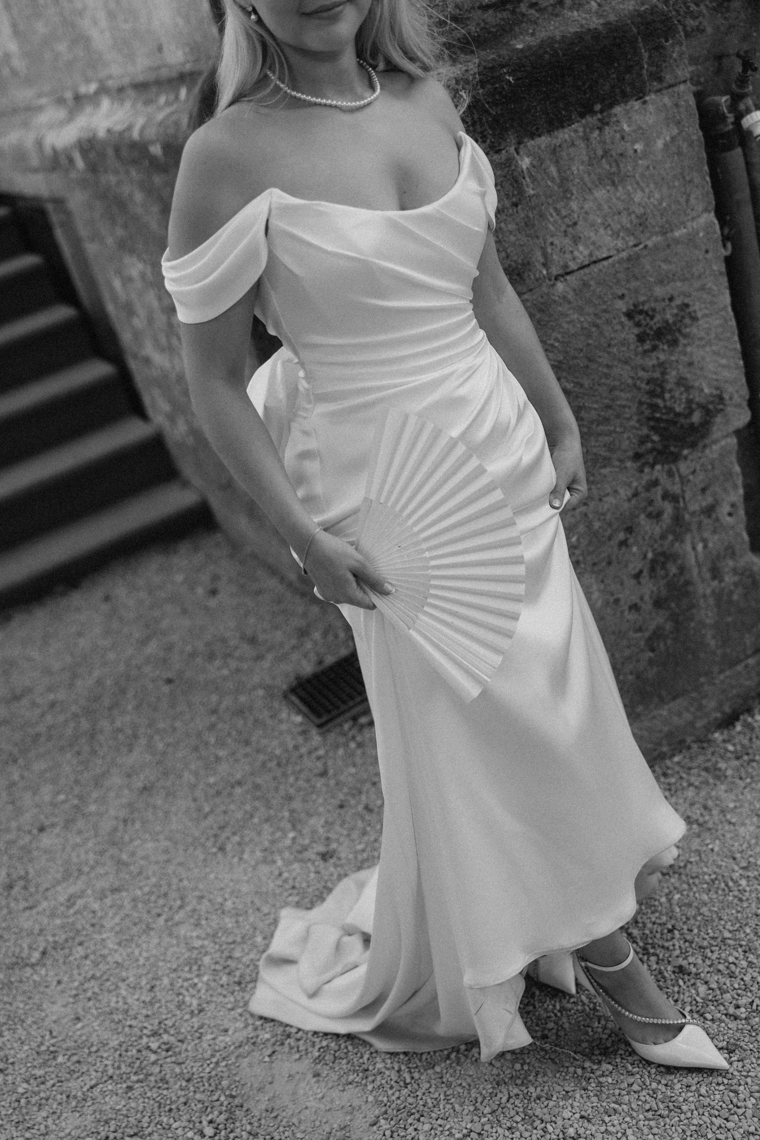 Bride in off-shoulder white satin gown holding a fan, wearing heels, black and white photo.