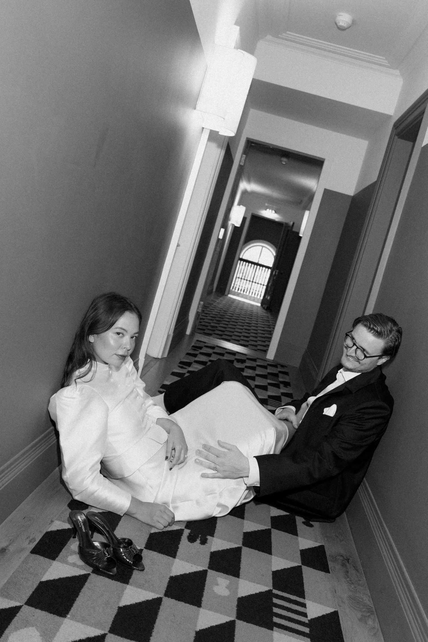 Black and white photo of a bride and groom sitting on a patterned hallway floor, bride in white gown, groom in dark suit.