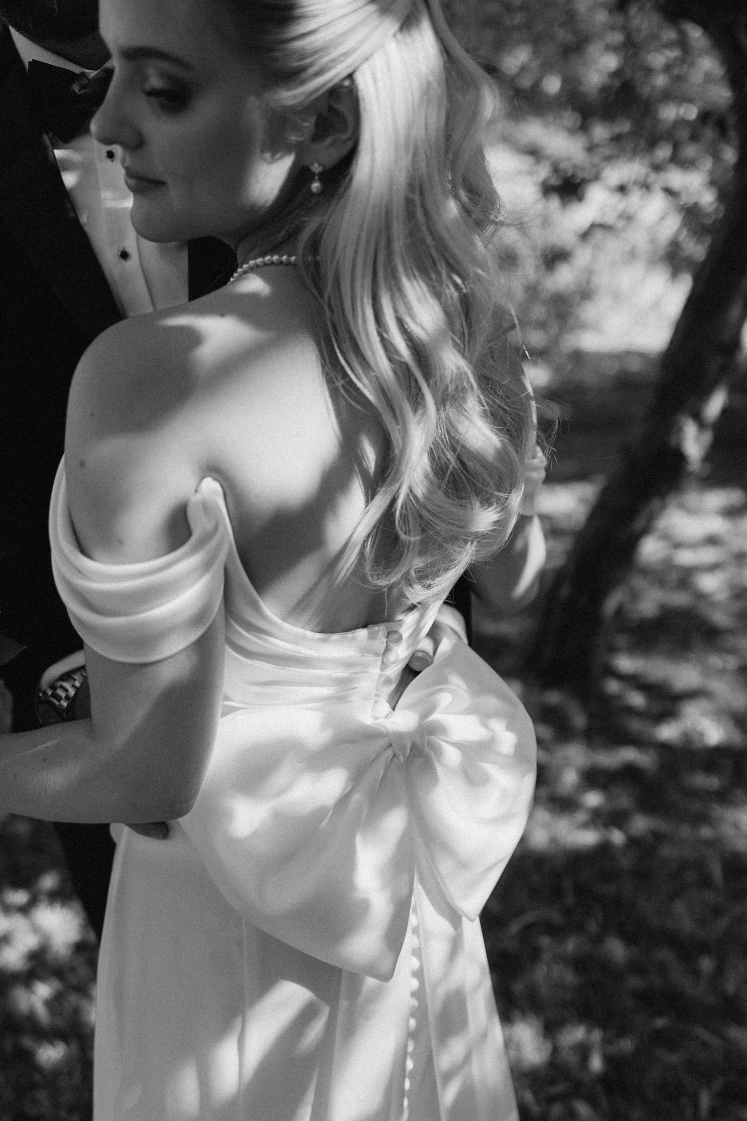 Black and white photo of a bride in an off-shoulder gown with a bow, embraced by groom outdoors.