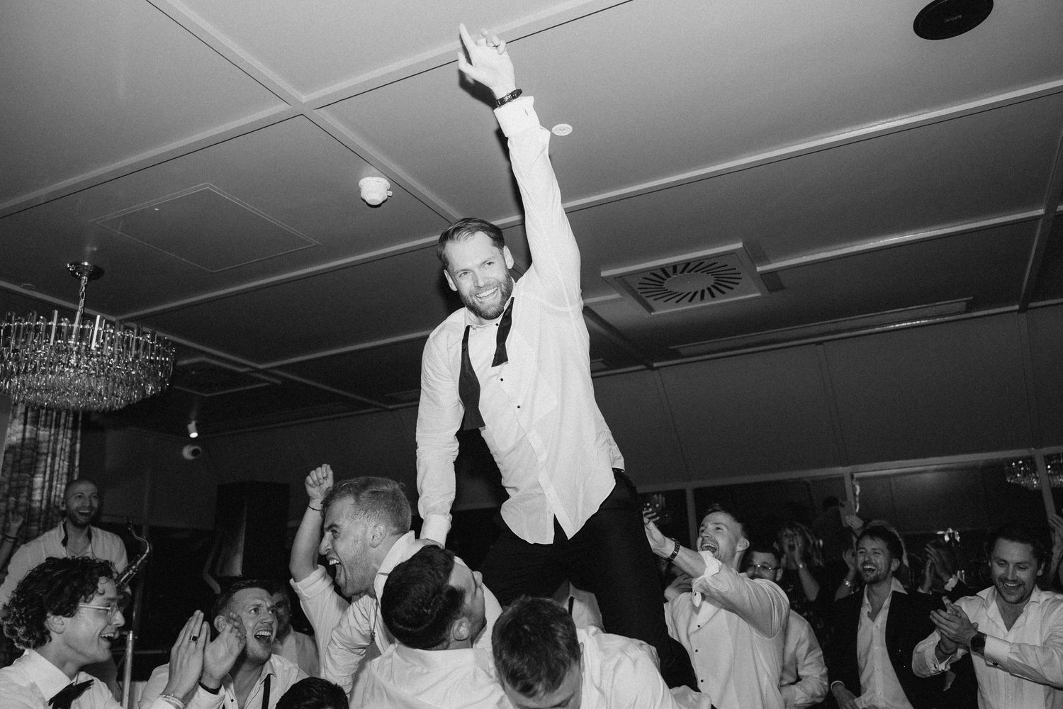 Man lifted by crowd at wedding reception celebration, black and white photo with chandelier backdrop.