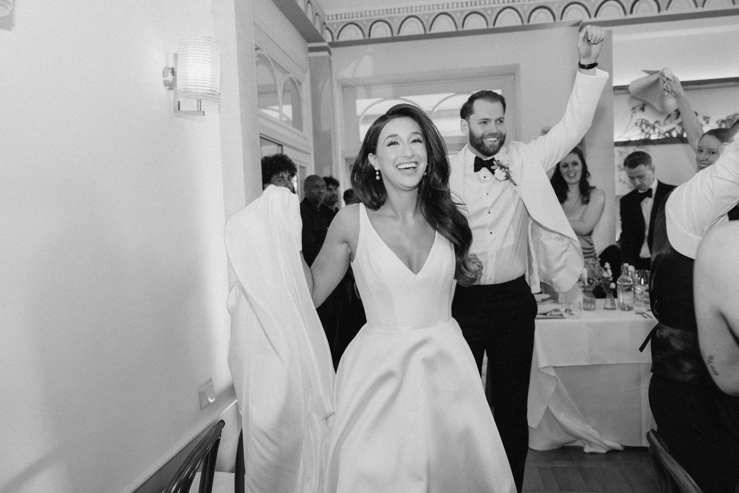 Black and white photo of a smiling bride in a white gown entering her wedding reception with the groom cheering behind her.