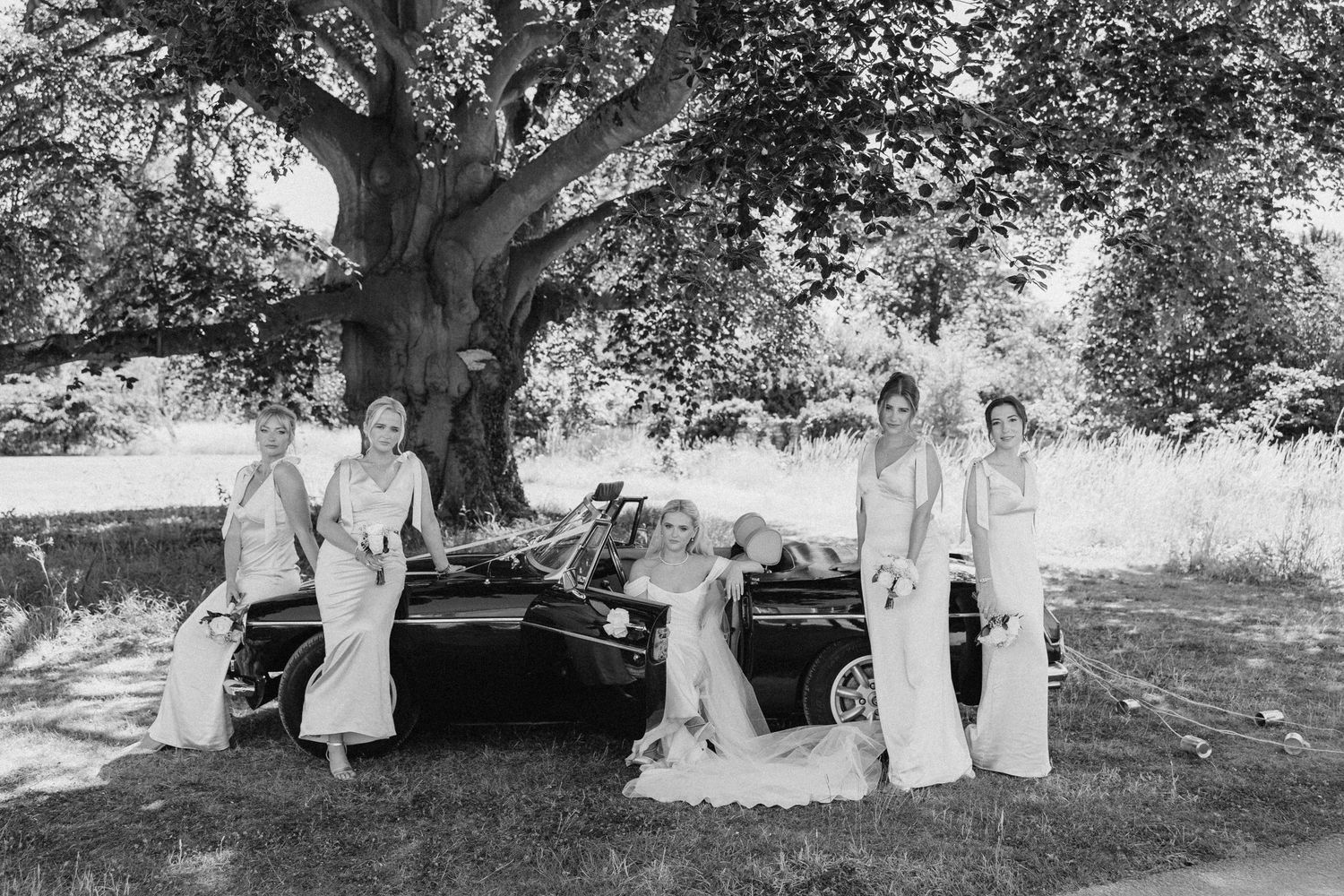 Black and white photo of bridesmaids and bride posing by a vintage car under a large tree.