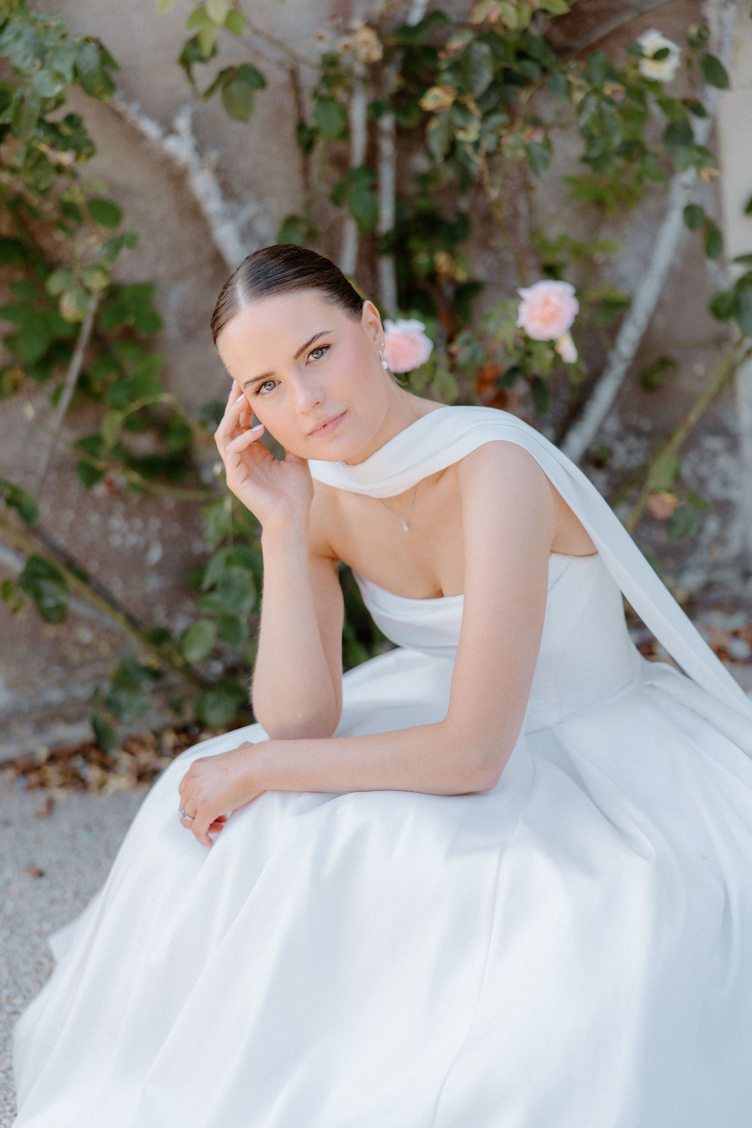 Elegant bride in white strapless gown with draped detail sits gracefully outdoors near roses.