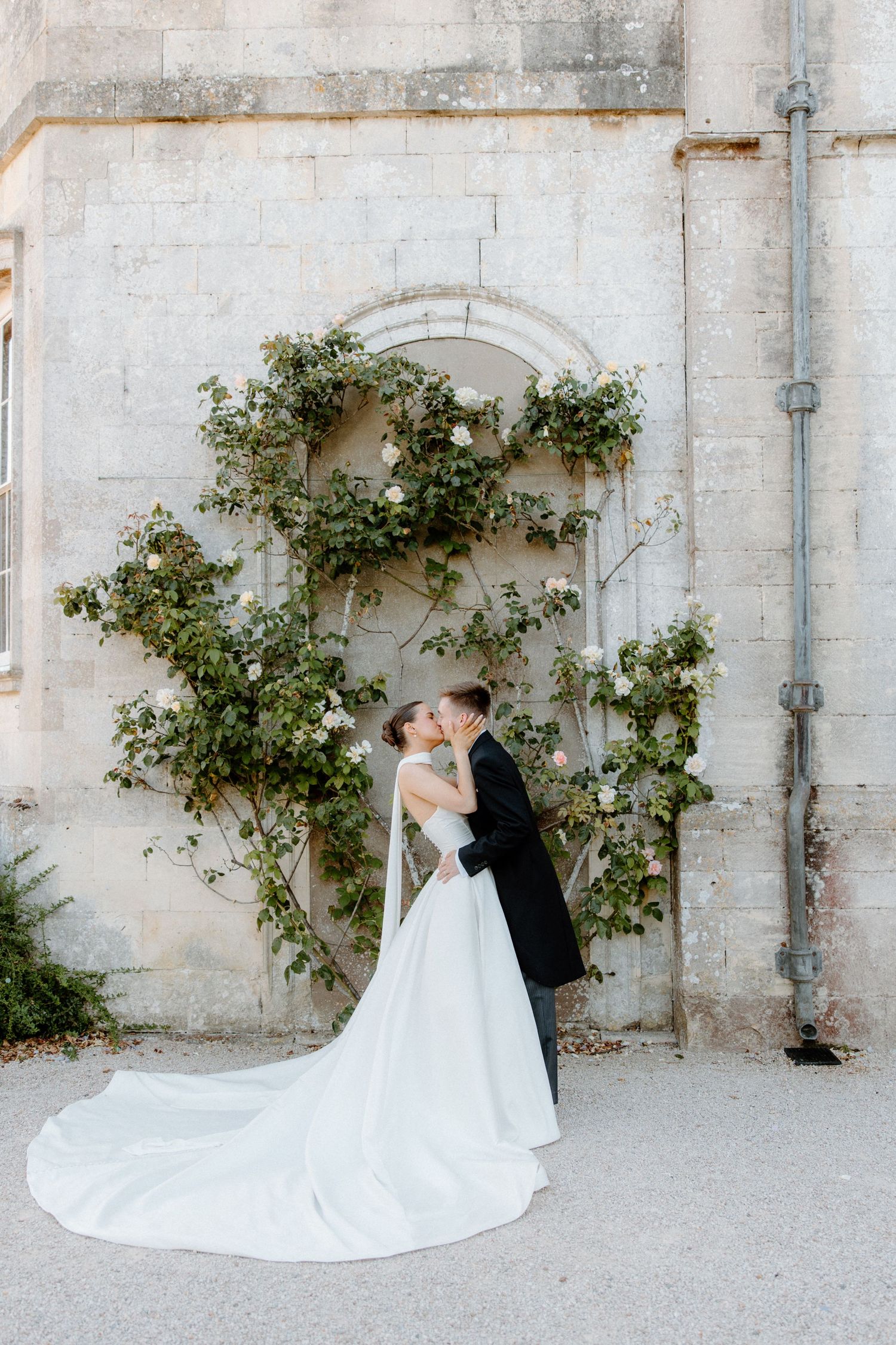 A bride and groom share a kiss in front of a stone wall adorned with climbing roses at their elegant wedding.