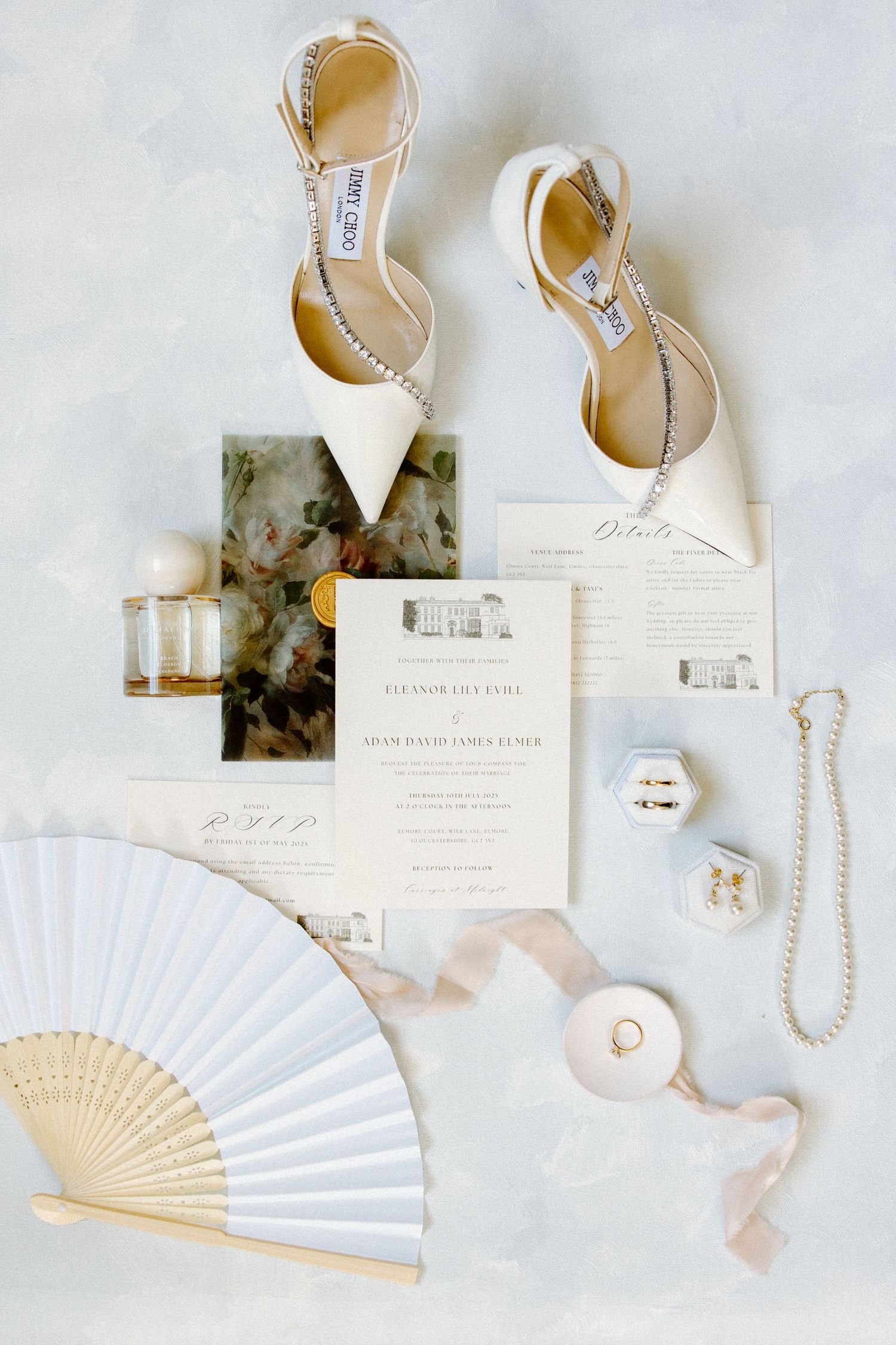 Bridal flat lay with Jimmy Choo heels, wedding invitations, hand fan, perfume, and delicate jewelry on a soft gray surface.