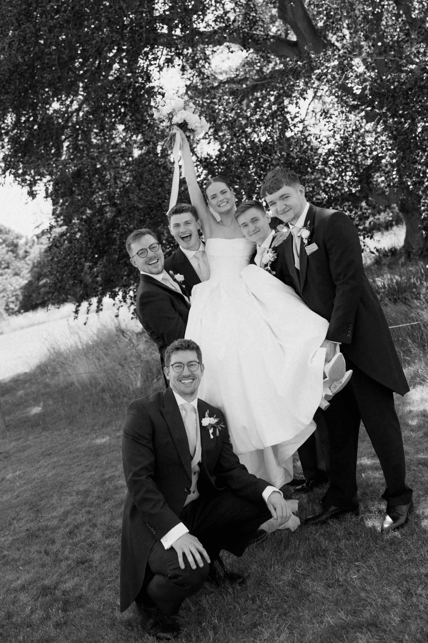 Black and white wedding photo of bride lifted by groomsmen outdoors under a tree.