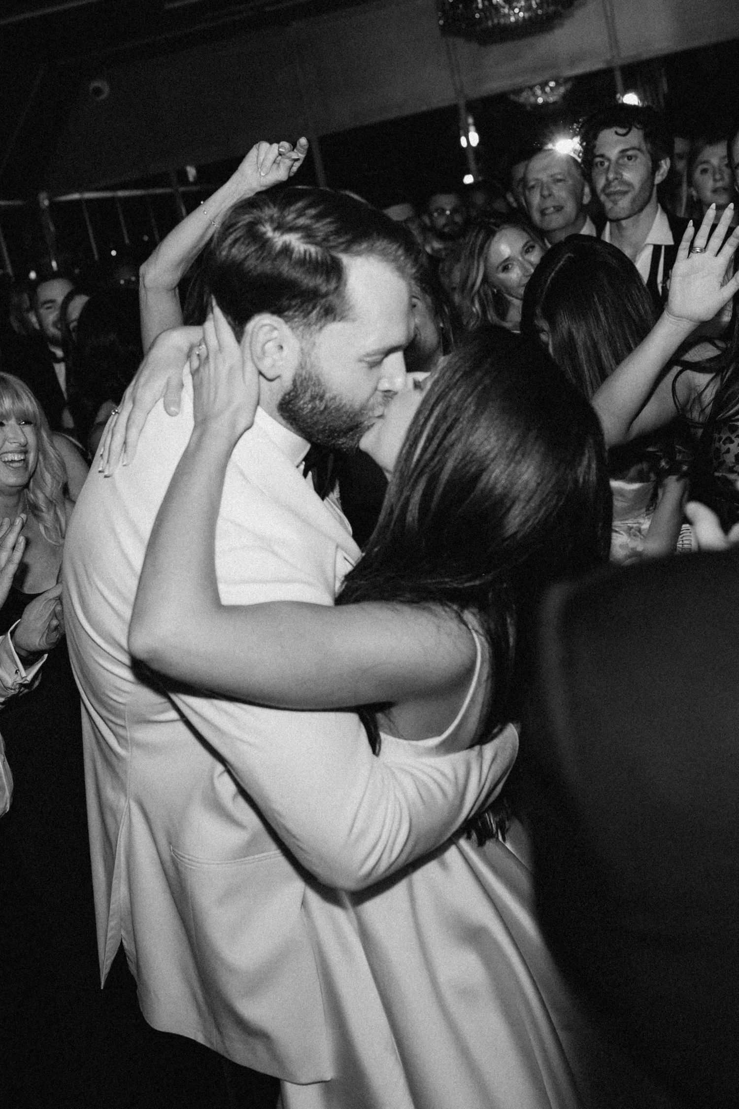 Black and white photo of a couple sharing a romantic kiss on a crowded dance floor at a wedding reception.
