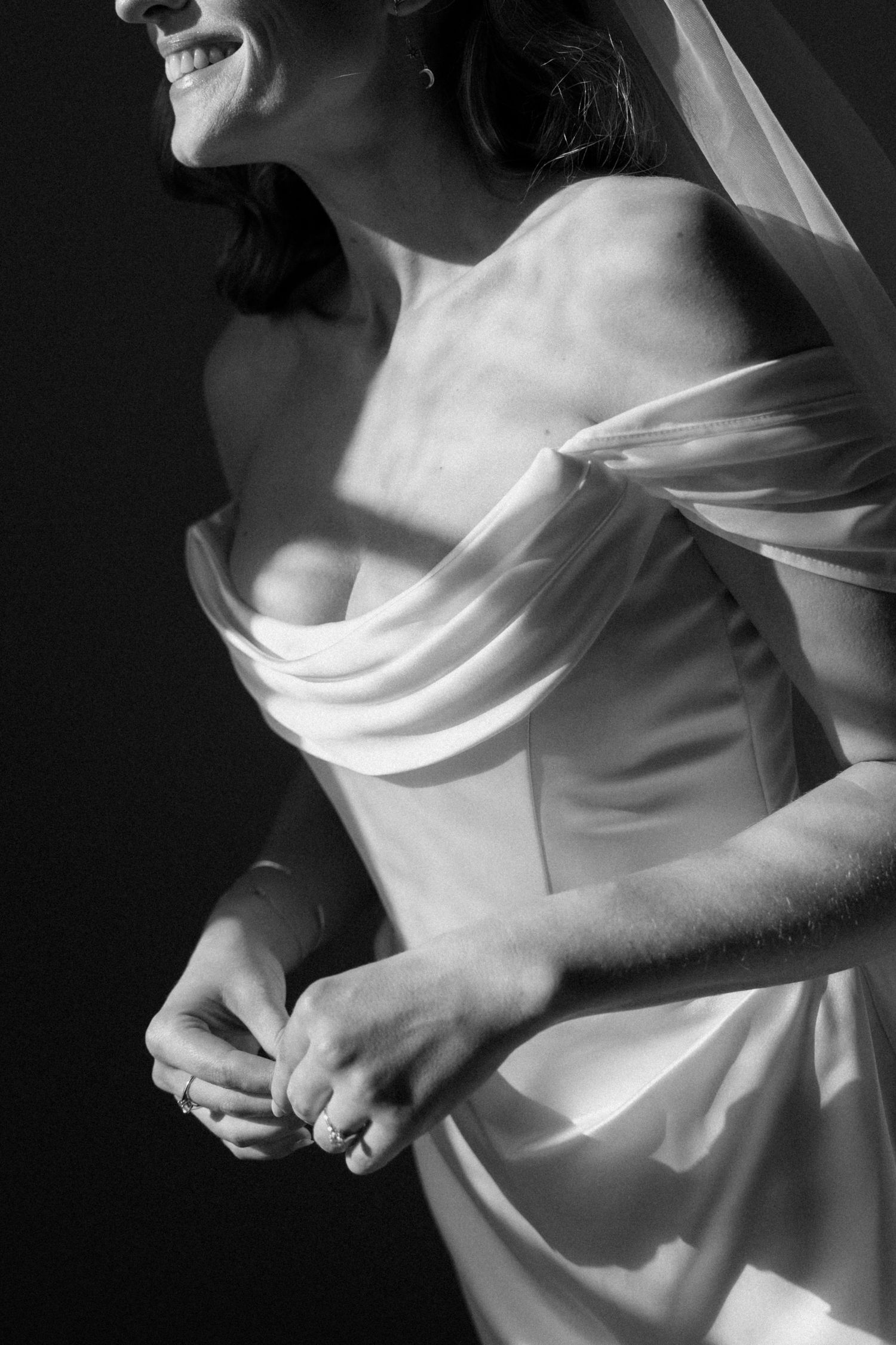 Black and white photo of a smiling bride in an off-shoulder wedding gown and veil, adjusting her ring.