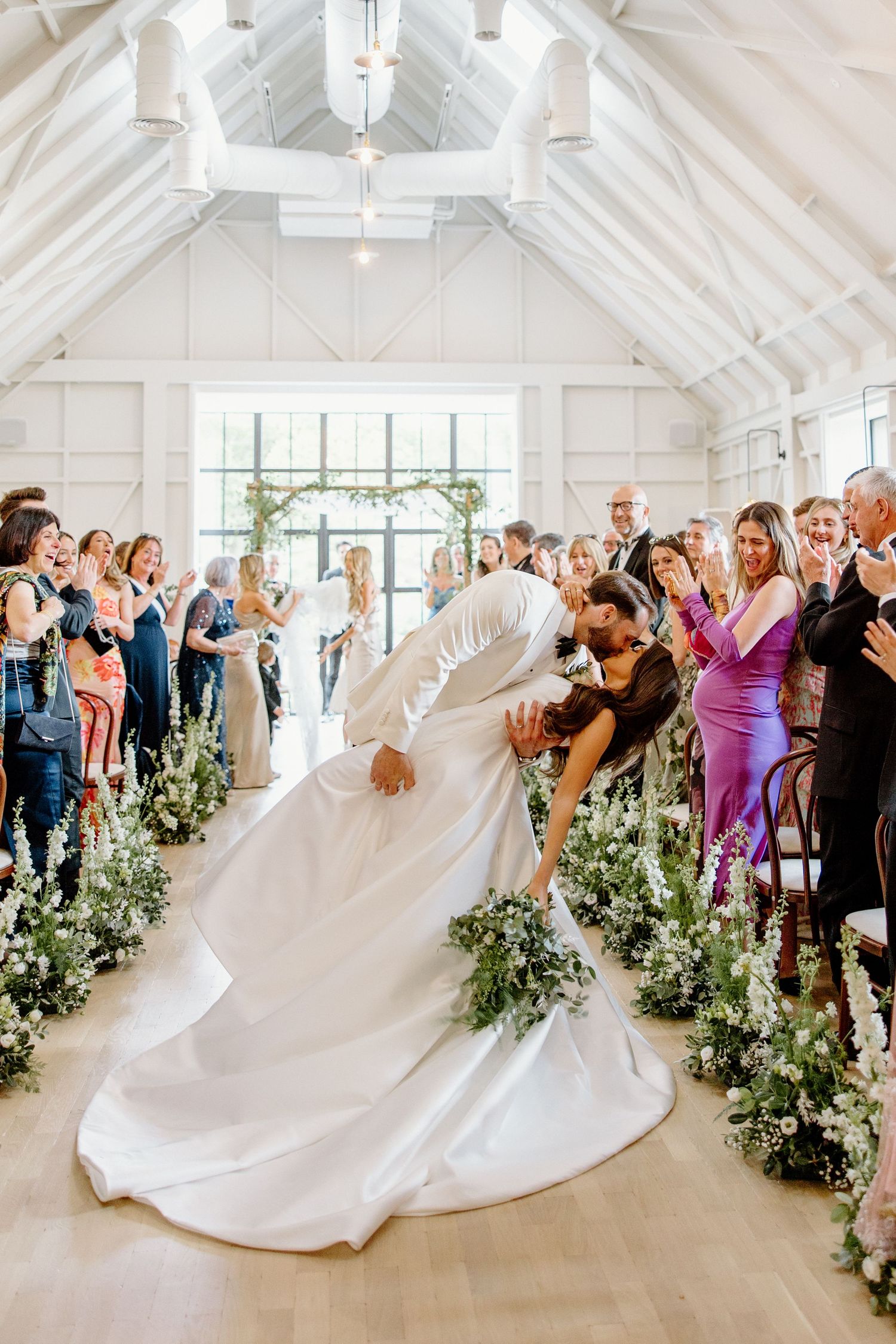 Bride and groom share a romantic dip kiss in the aisle of a bright white chapel wedding venue with floral arrangements.