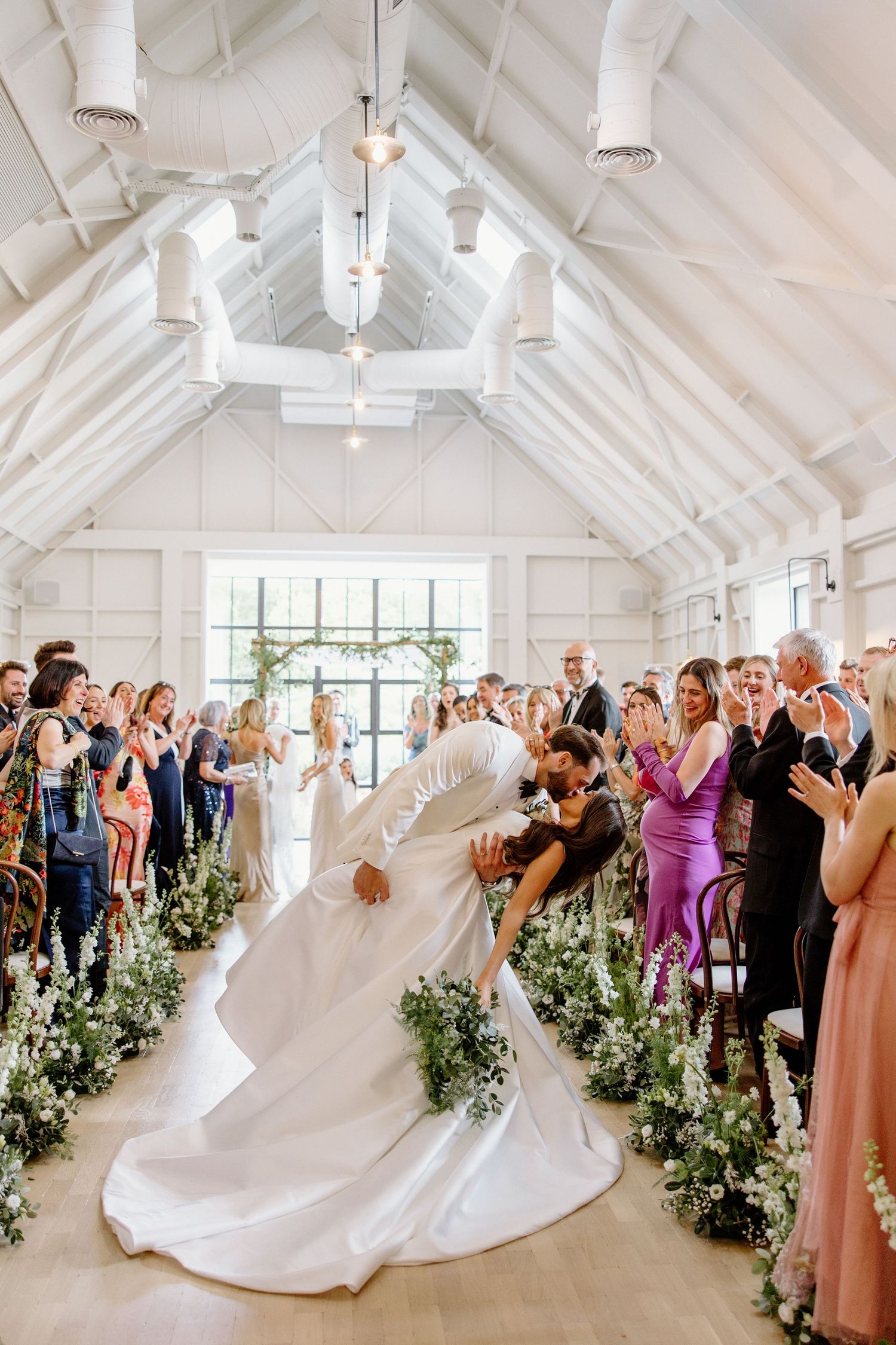 Bride and groom share a dip kiss at the altar in a bright white barn wedding venue lined with greenery.