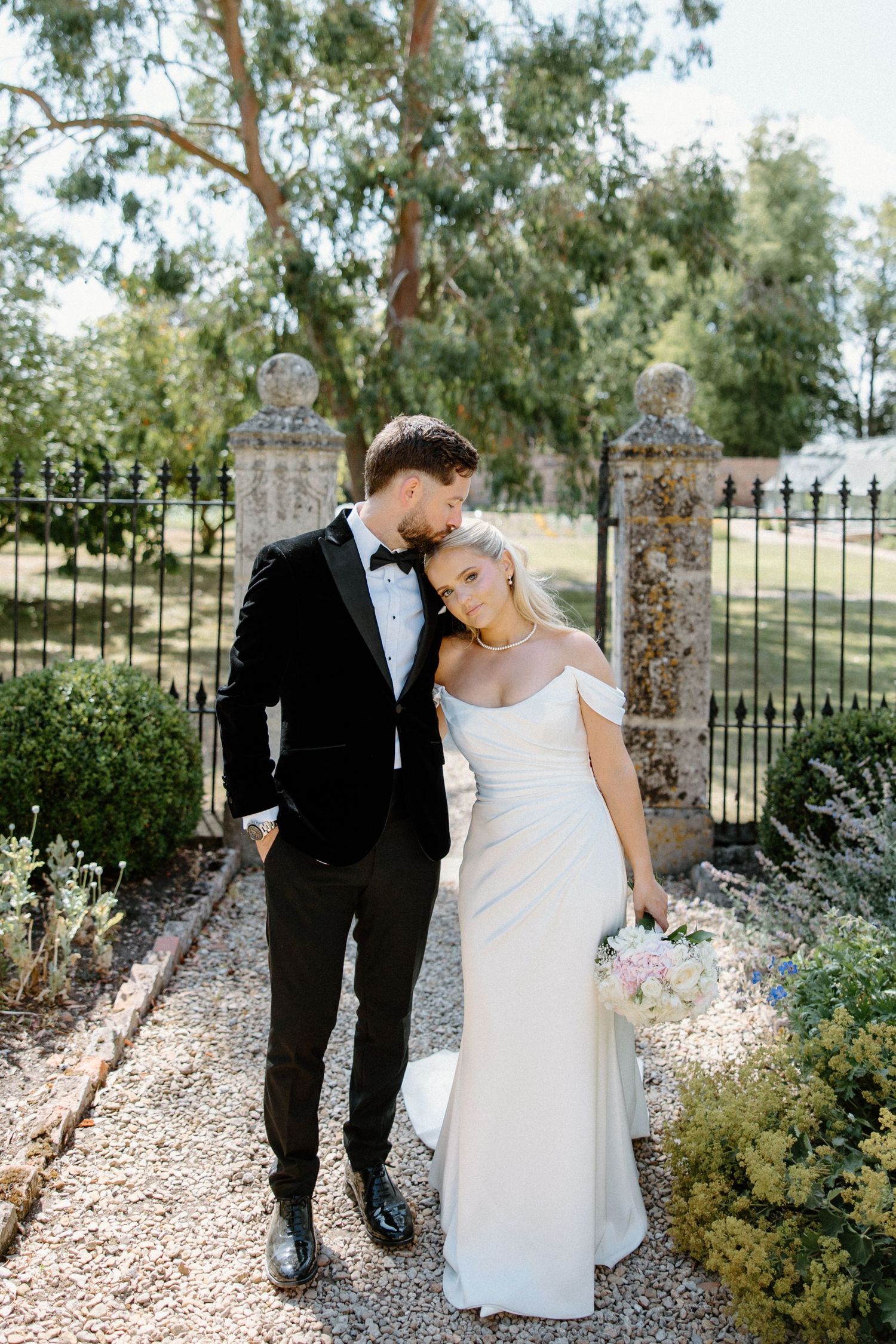Bride and groom pose on garden path, she in off-shoulder white gown, he in black tuxedo, holding bouquet.