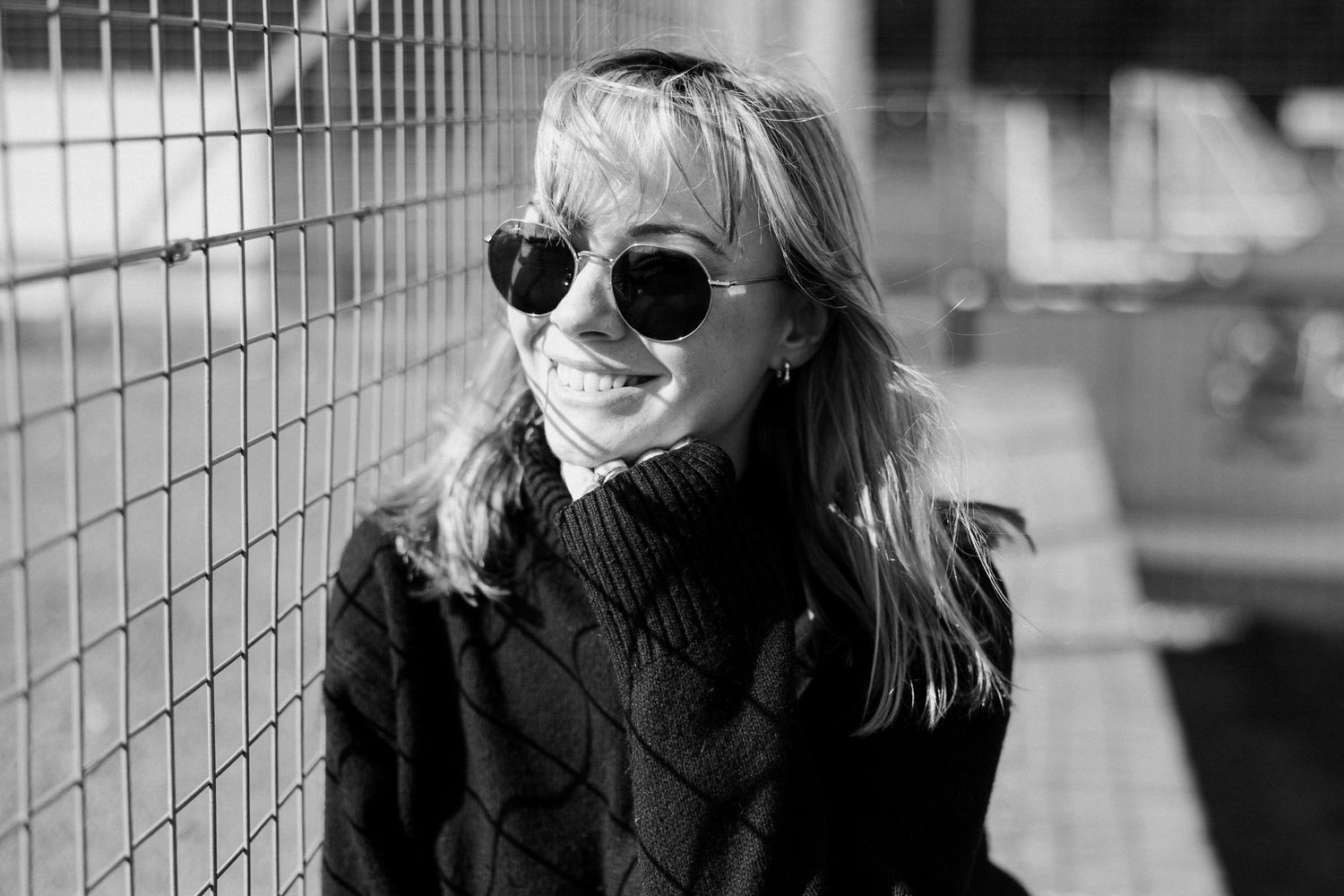 A black and white portrait of someone in sunglasses and a turtleneck sweater smiling while leaning against a fence.