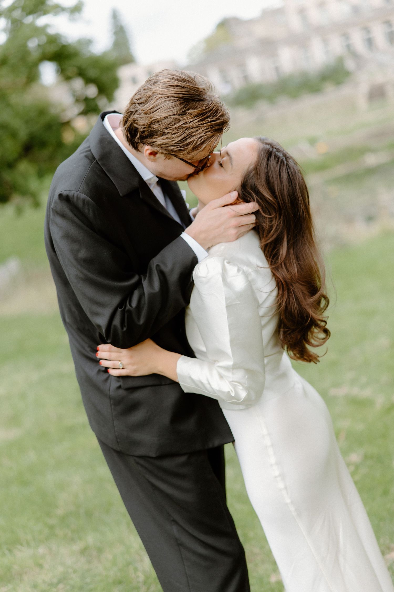A couple shares a romantic kiss outdoors, bride in white satin gown and groom in black suit on their wedding day.