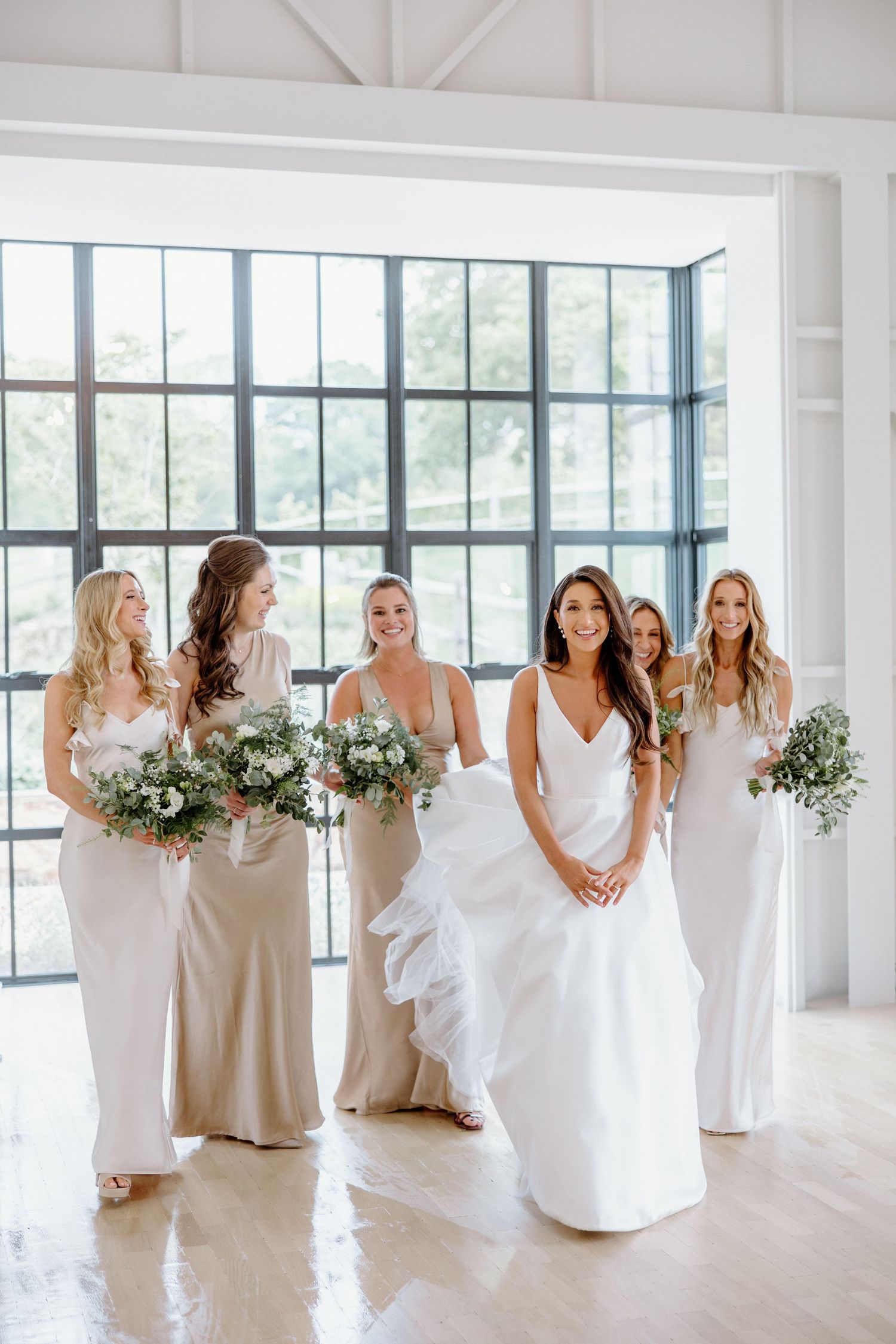 Bride in white gown holds dress while smiling with bridesmaids in champagne dresses holding greenery bouquets indoors.