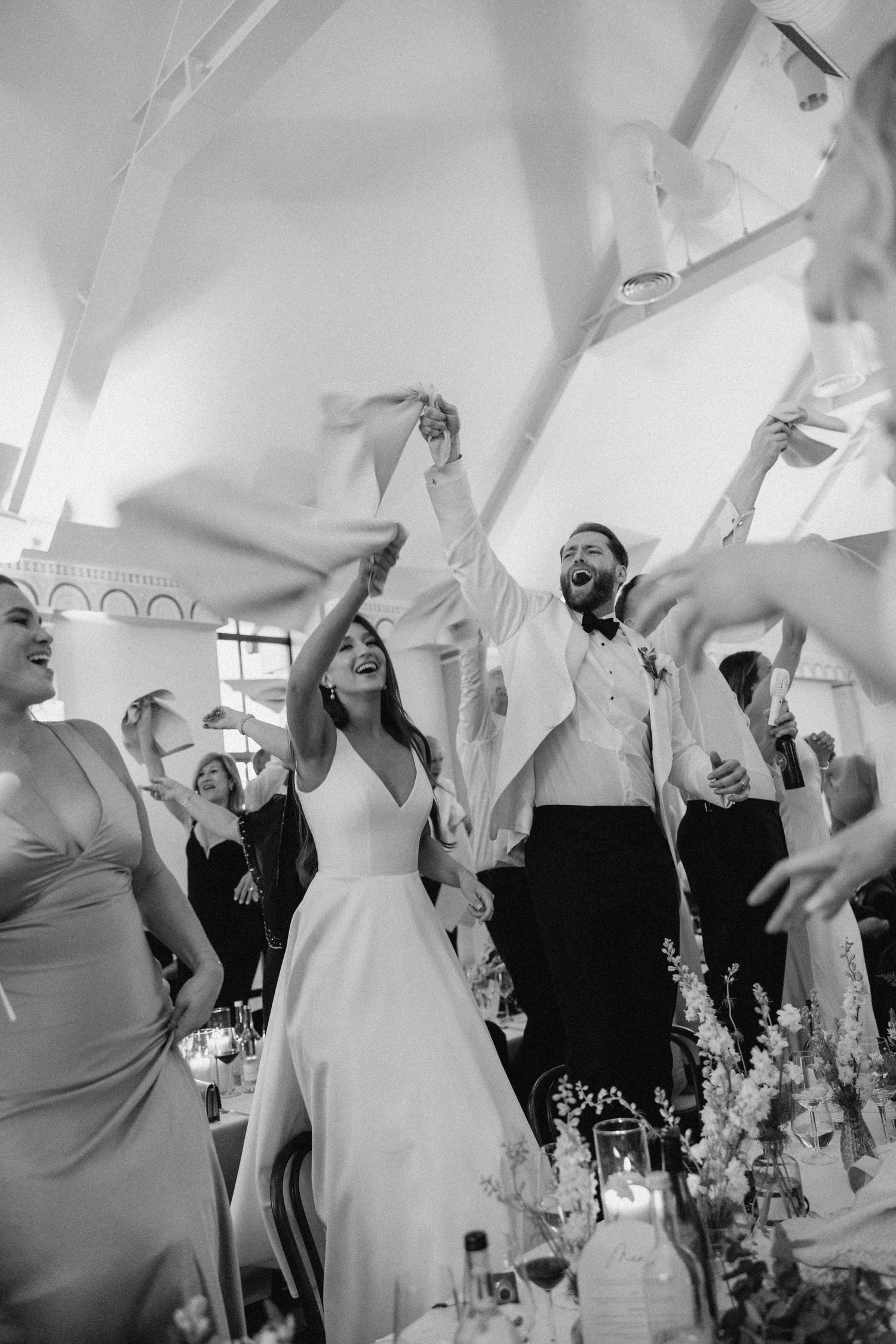 Black and white photo of joyful bride and groom celebrating their wedding reception with guests waving napkins.