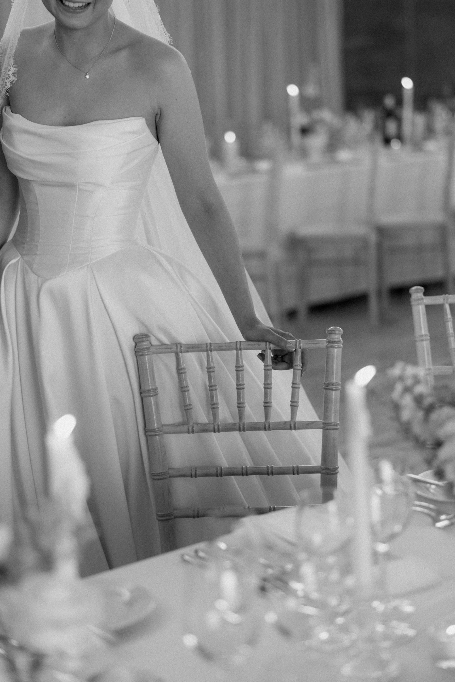 Black and white photo of a bride in a strapless gown standing beside a Chiavari chair at an elegant wedding reception.