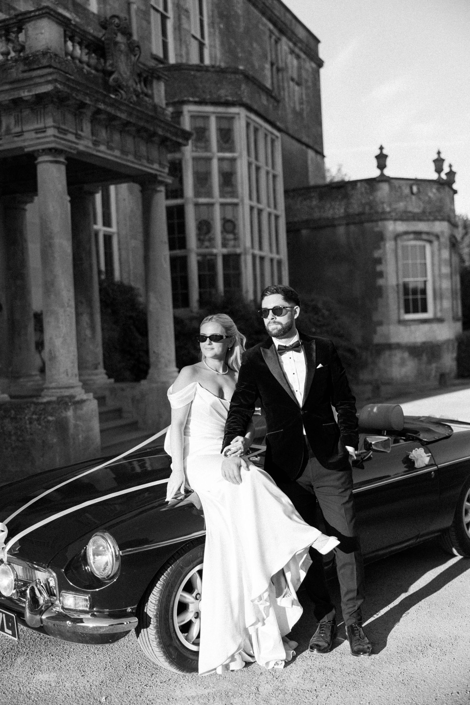 Black and white wedding photos showing couple in formal attire posing by luxury car in elegant architectural setting.