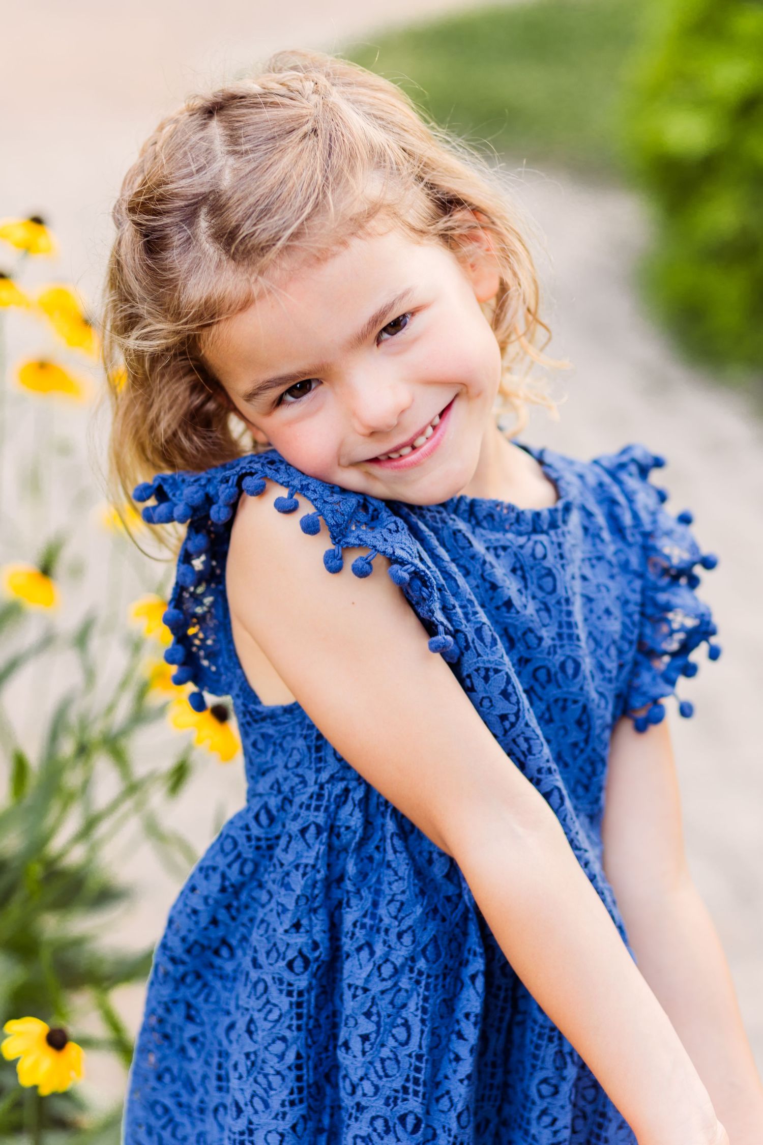 Young child in blue pom-pom dress smiling near yellow flowers outdoors.