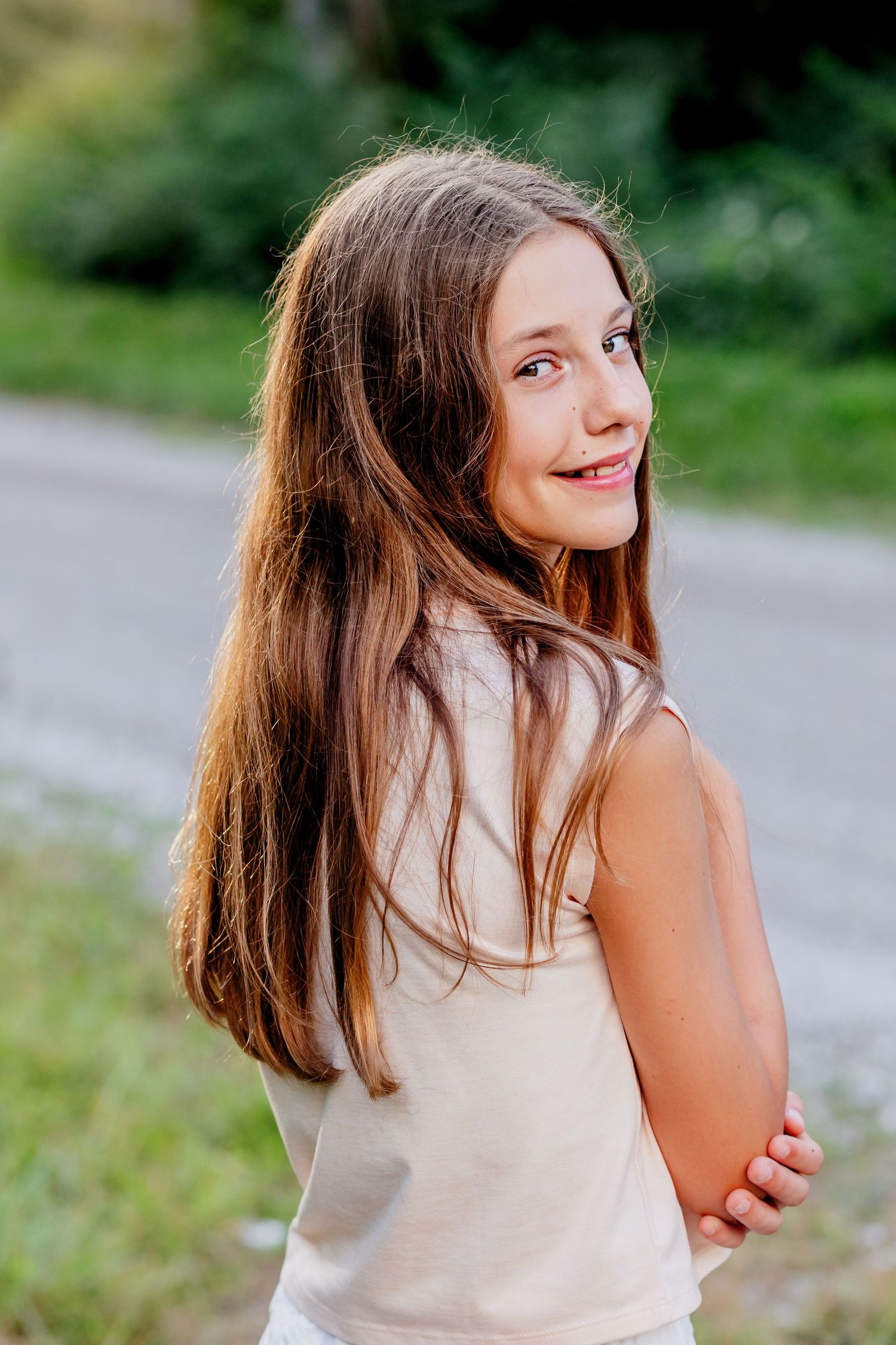 Outdoor portrait of someone with long brown hair smiling over their shoulder.