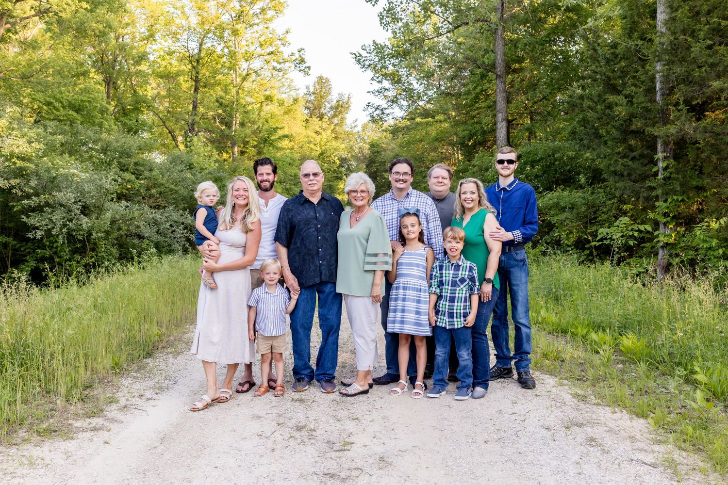 Multi-generational family group portrait on a dirt path surrounded by green summer foliage.