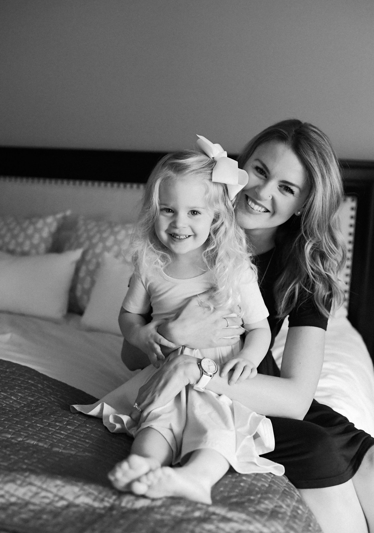 Black and white photo of mother and young daughter with blonde hair and hair bow sitting together on a bed.