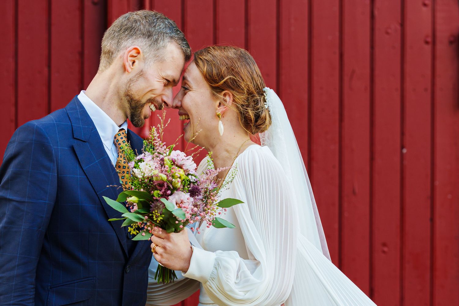 A happy couple shares an intimate moment on their wedding day against a red barn wall, with the bride holding a floral bouquet.