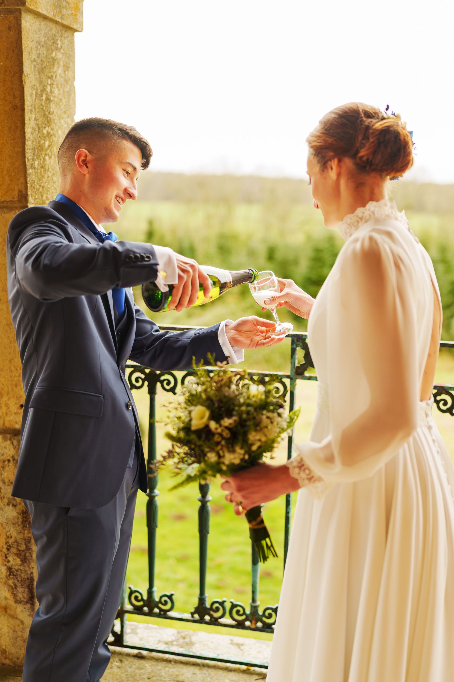 Couple sharing champagne during wedding celebration on outdoor porch with wrought iron railings and green landscape.