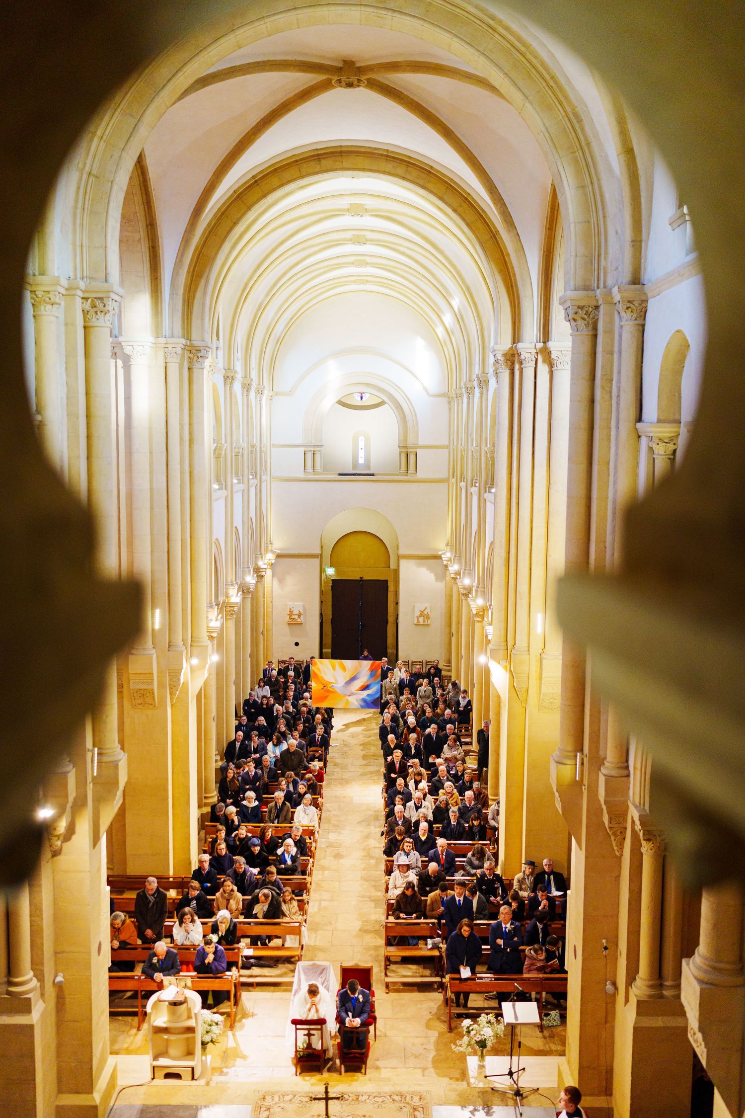Elevated view of an ornate church interior with vaulted arches and rows of people seated in pews during a ceremony.