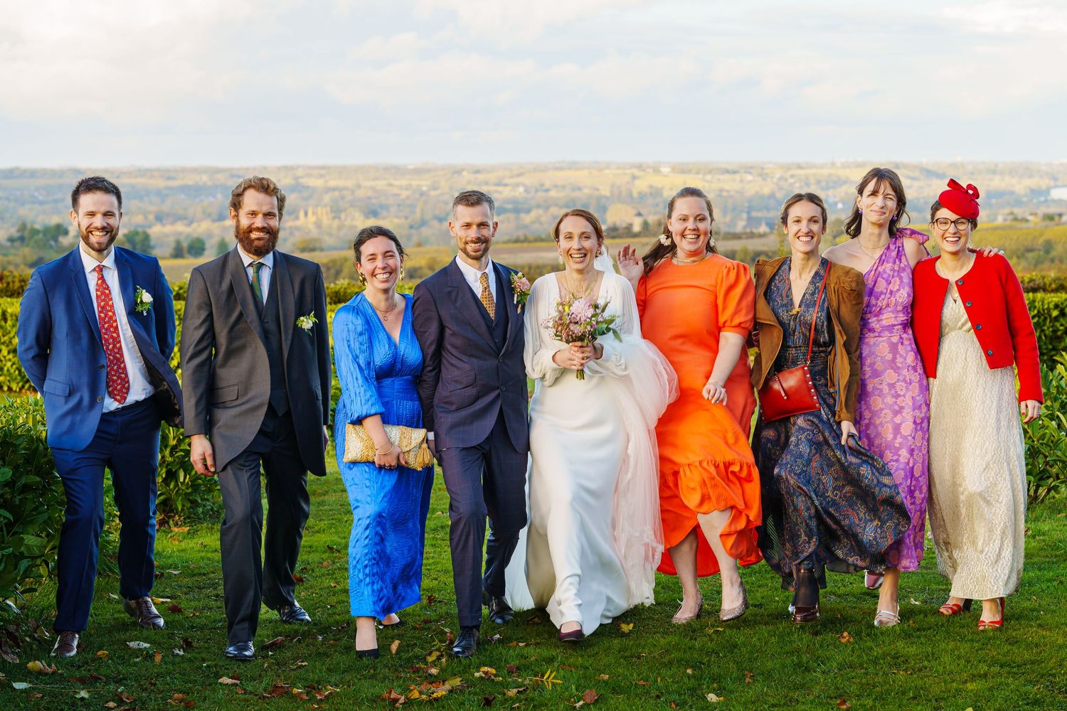 Wedding party lined up outdoors wearing vibrant mix of formal attire in blue, orange, and white against green landscape.