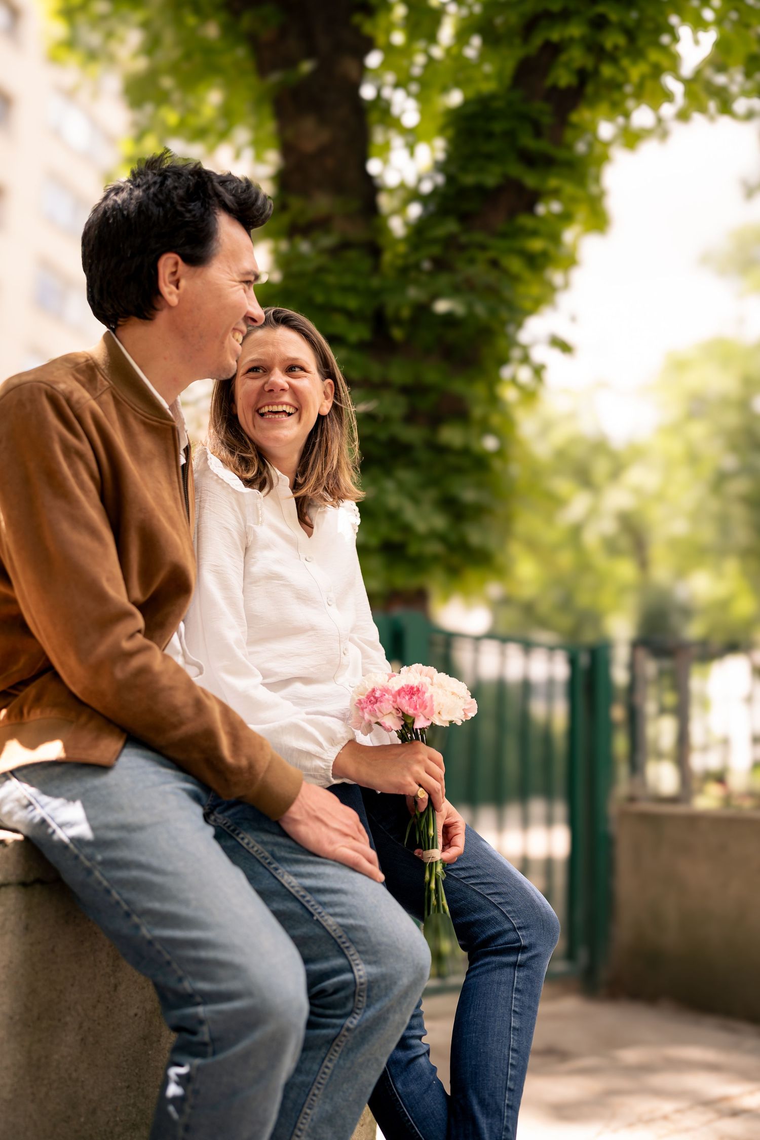 Couple laughing together while sitting on stone wall in park setting.