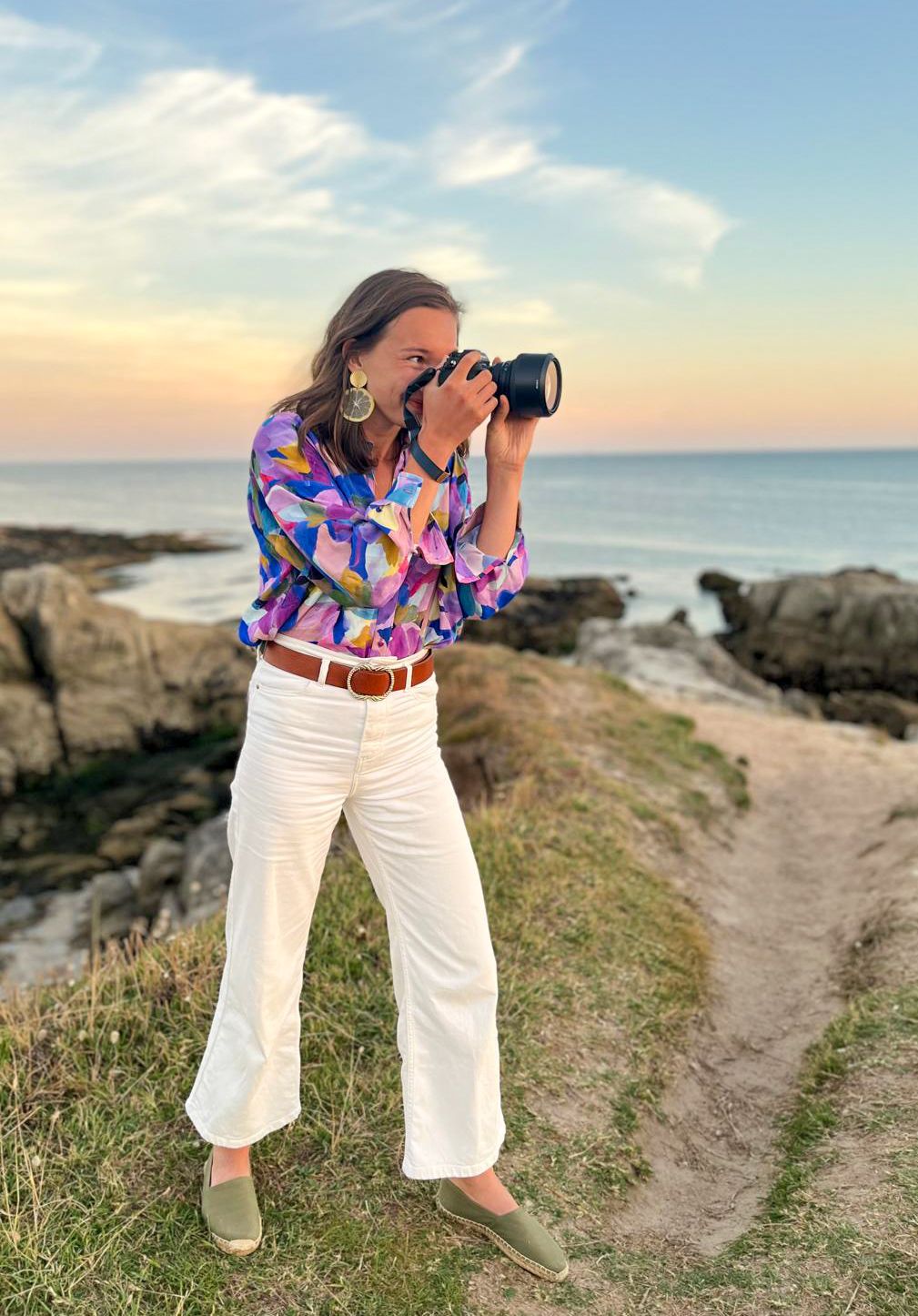 Photographer in colorful patterned shirt and white pants captures coastal scenery at sunset.