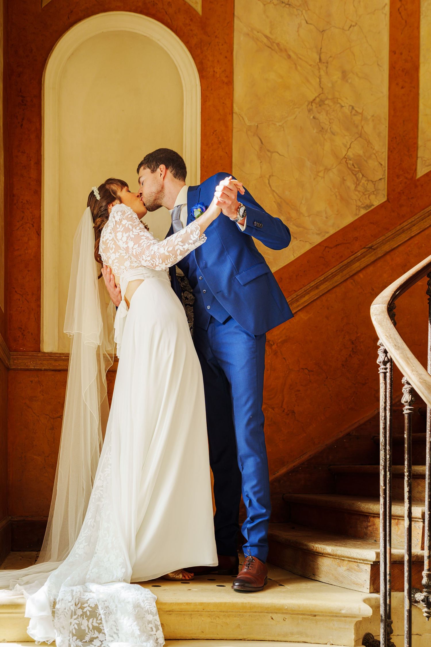 Romantic wedding couple in blue suit and white dress share intimate moments along an elegant marble staircase.