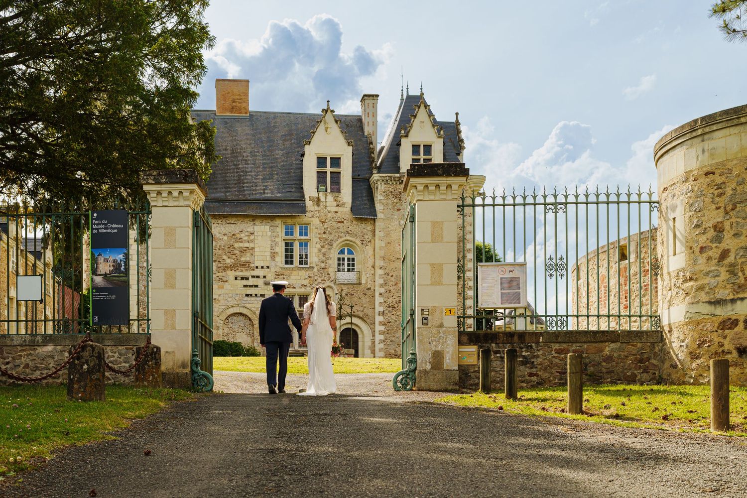 Bride and groom walking hand in hand toward an elegant French château gate surrounded by lush greenery.