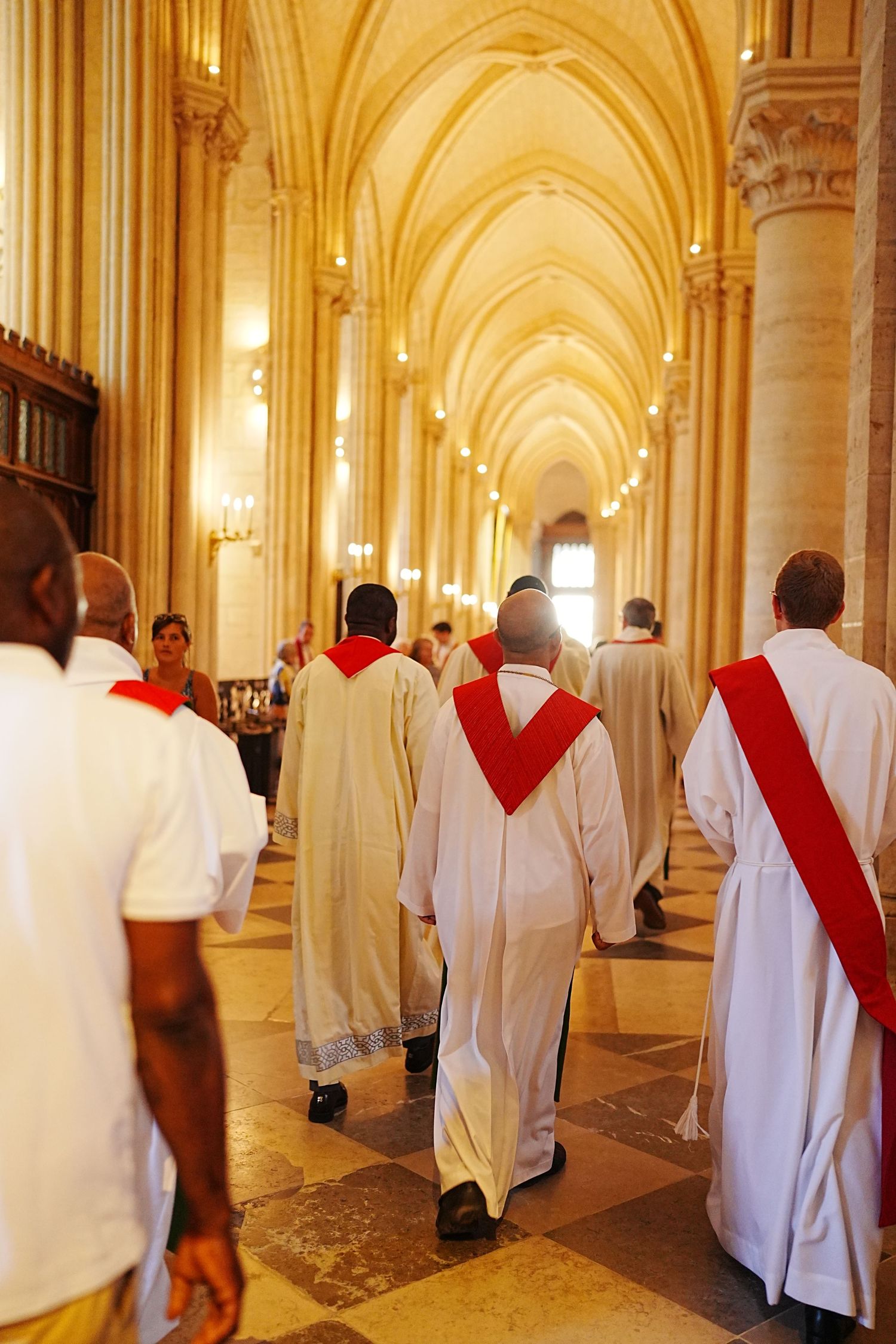 Clergy members in white robes and red stoles walking through cathedral hallway.