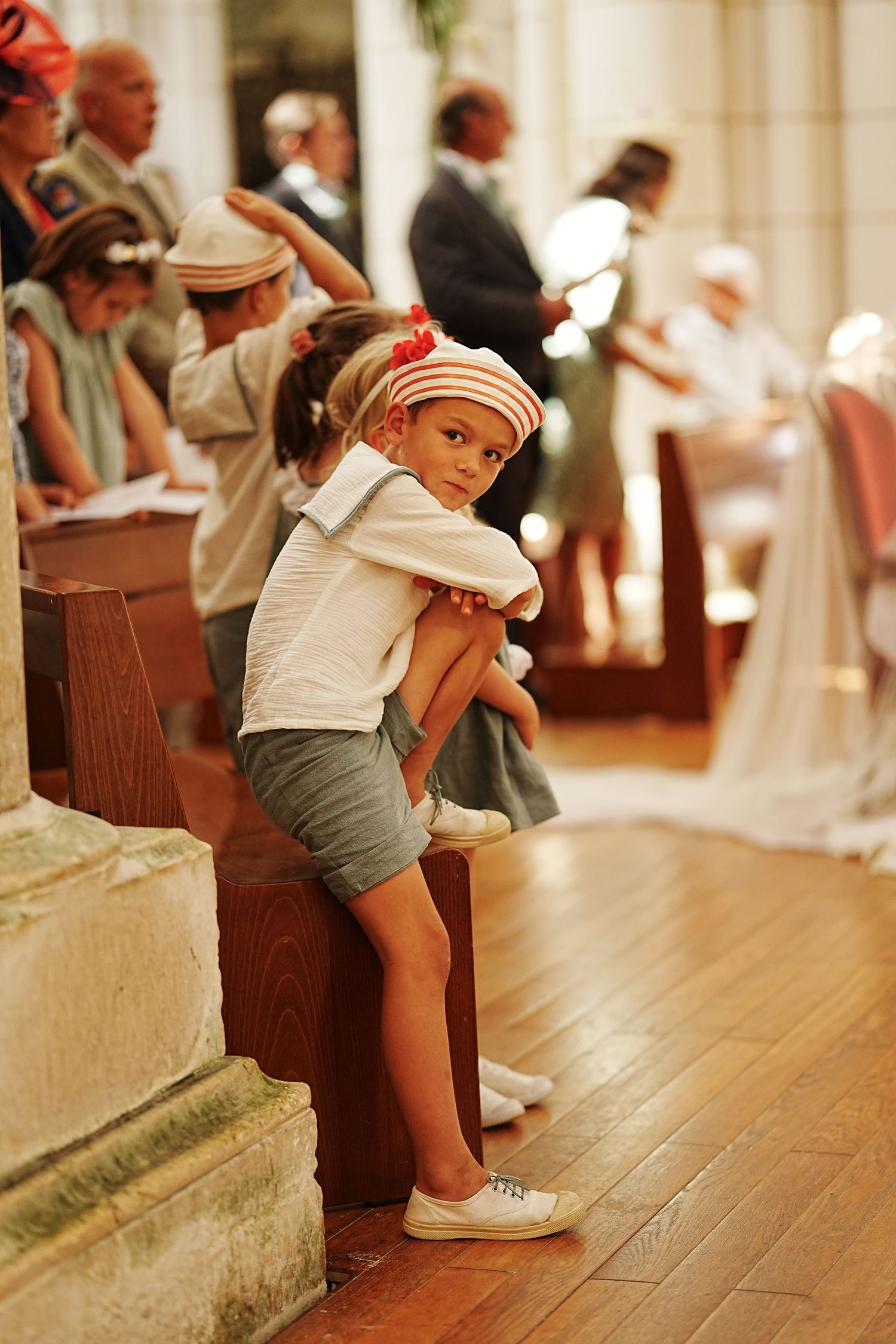 Child sitting on wooden church pew in casual summer attire.