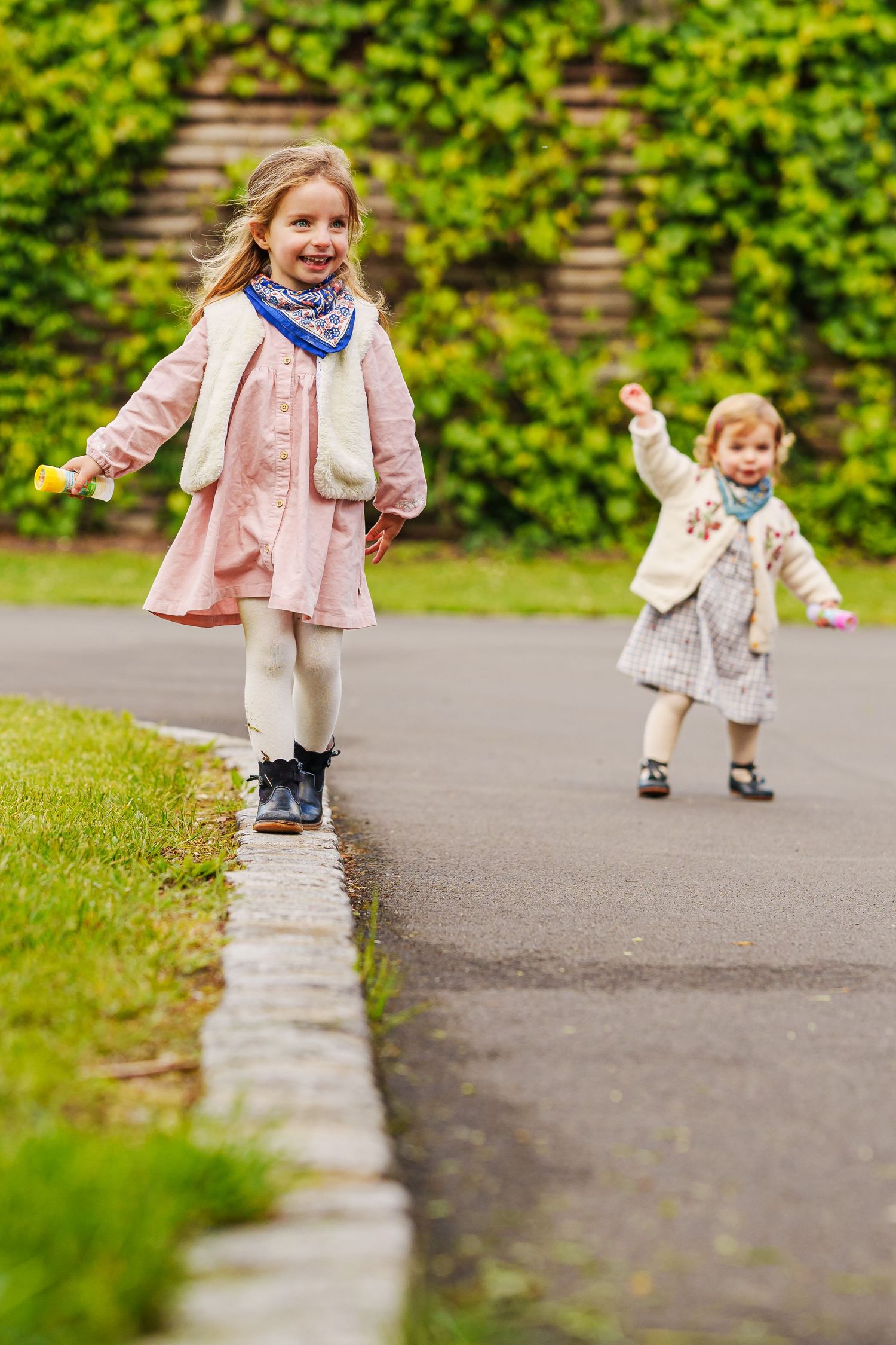 Two young girls in pink coats and scarves playing on a garden path with green ivy wall backdrop.