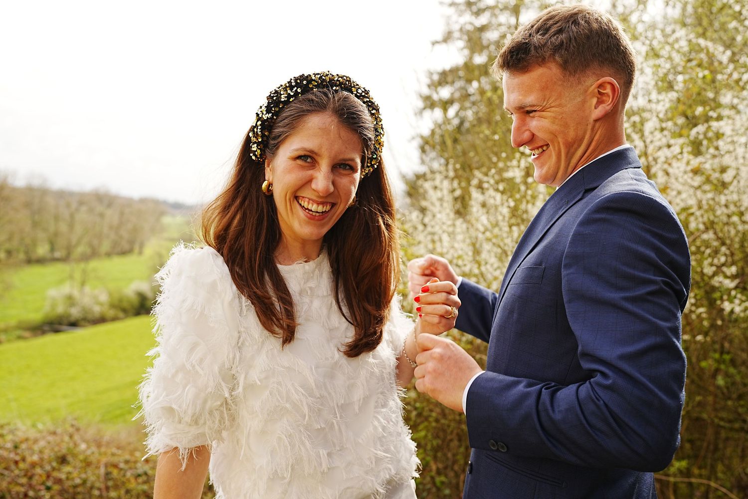 Happy couple in wedding attire laughing outdoors, bride in white feathered dress with floral headband, groom in navy suit.