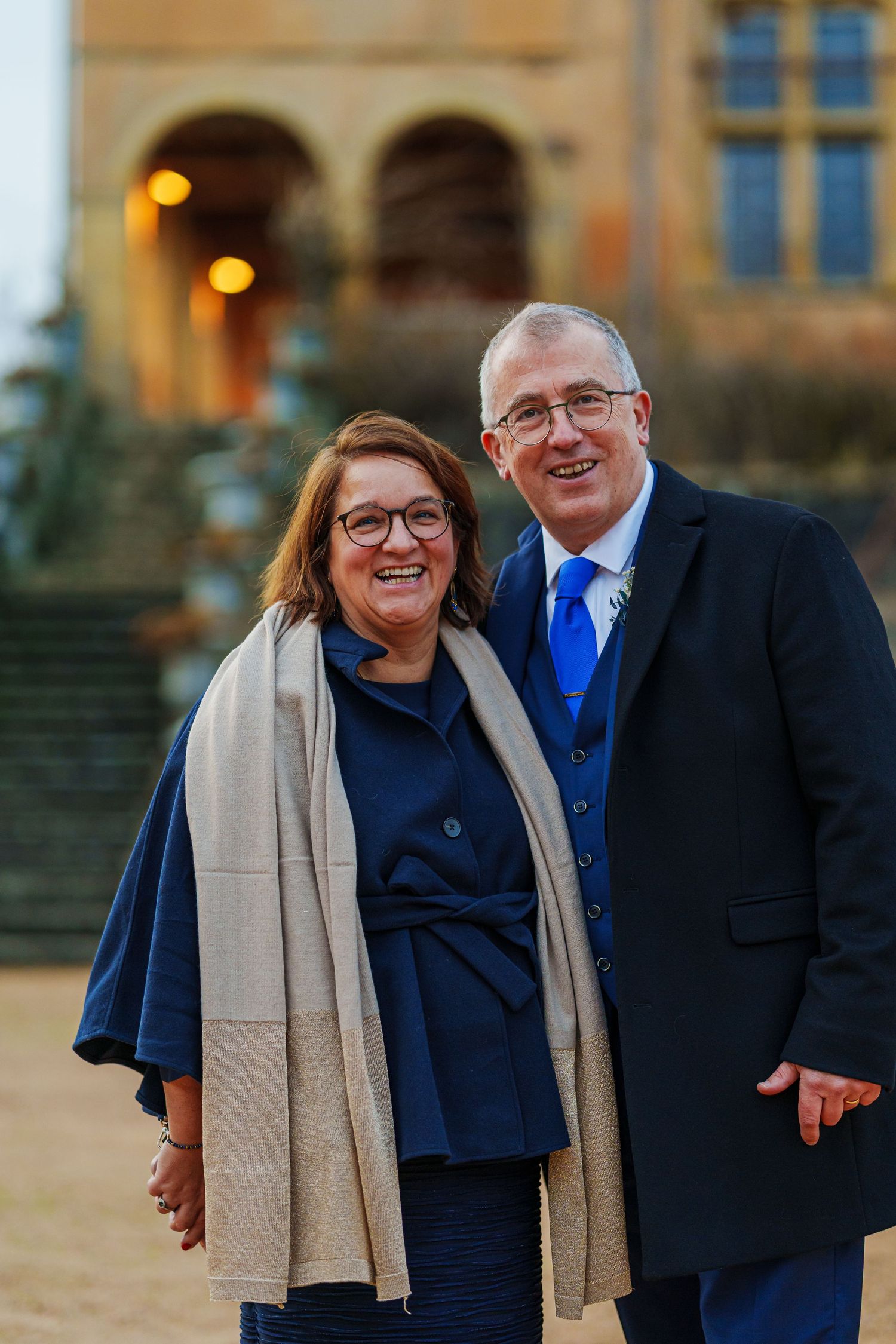 A couple smiles together outdoors during a photo session at golden hour with architectural elements in the background.