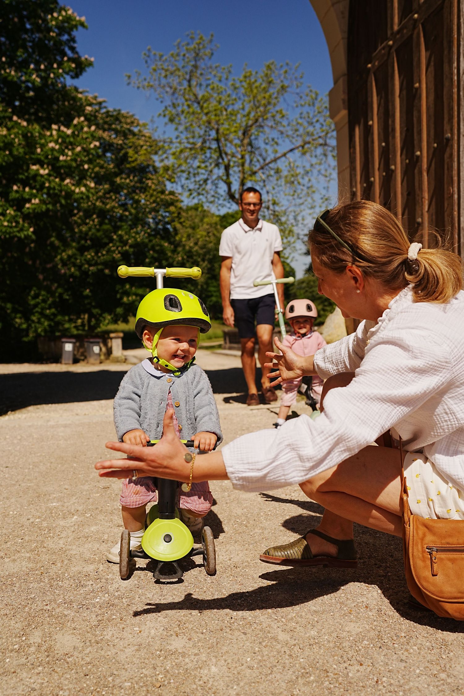 Child riding skateboard while adult crouches to take photo in sunny courtyard.