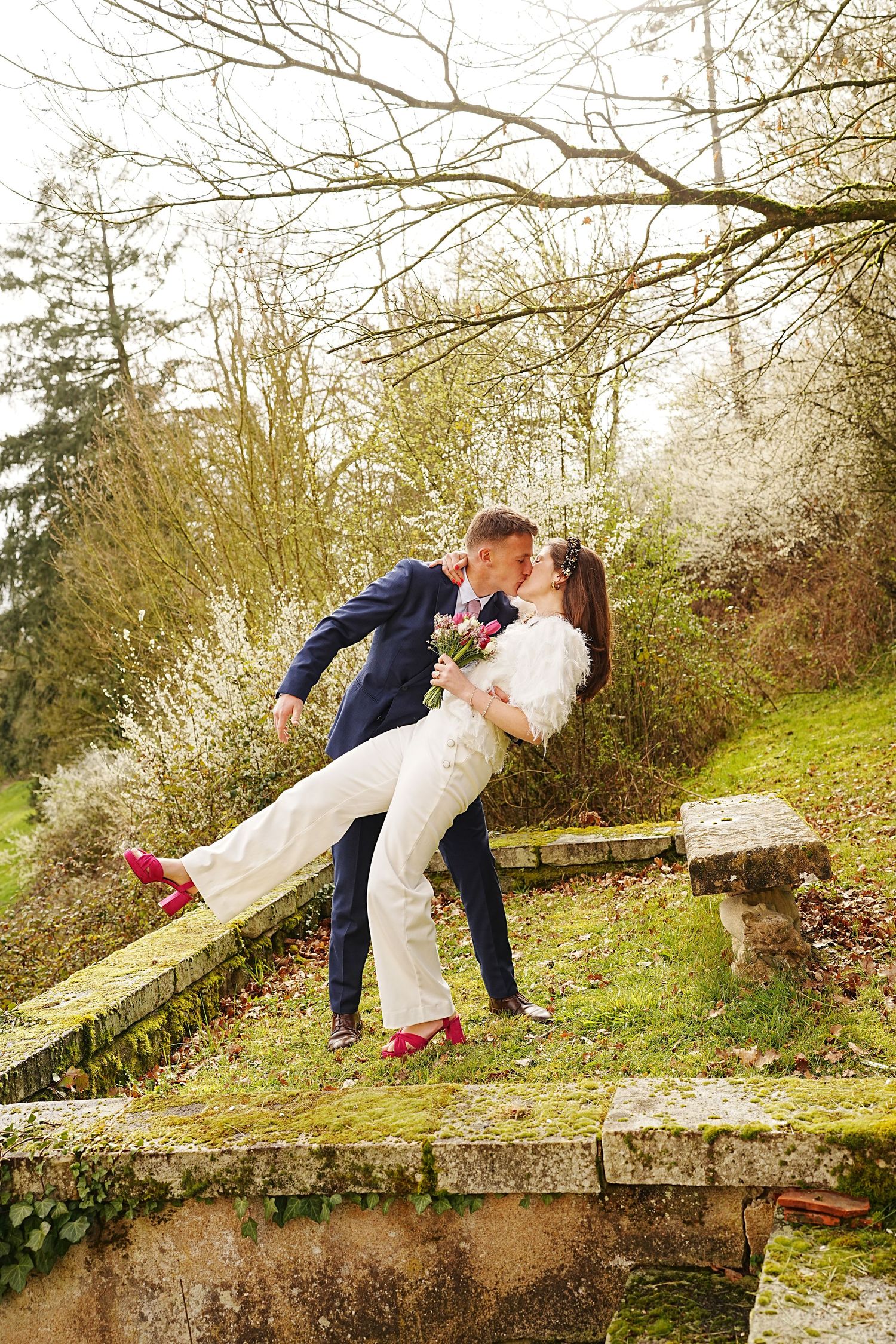 Bride in white jumpsuit and red heels shares a romantic dip kiss with groom in navy suit outdoors.
